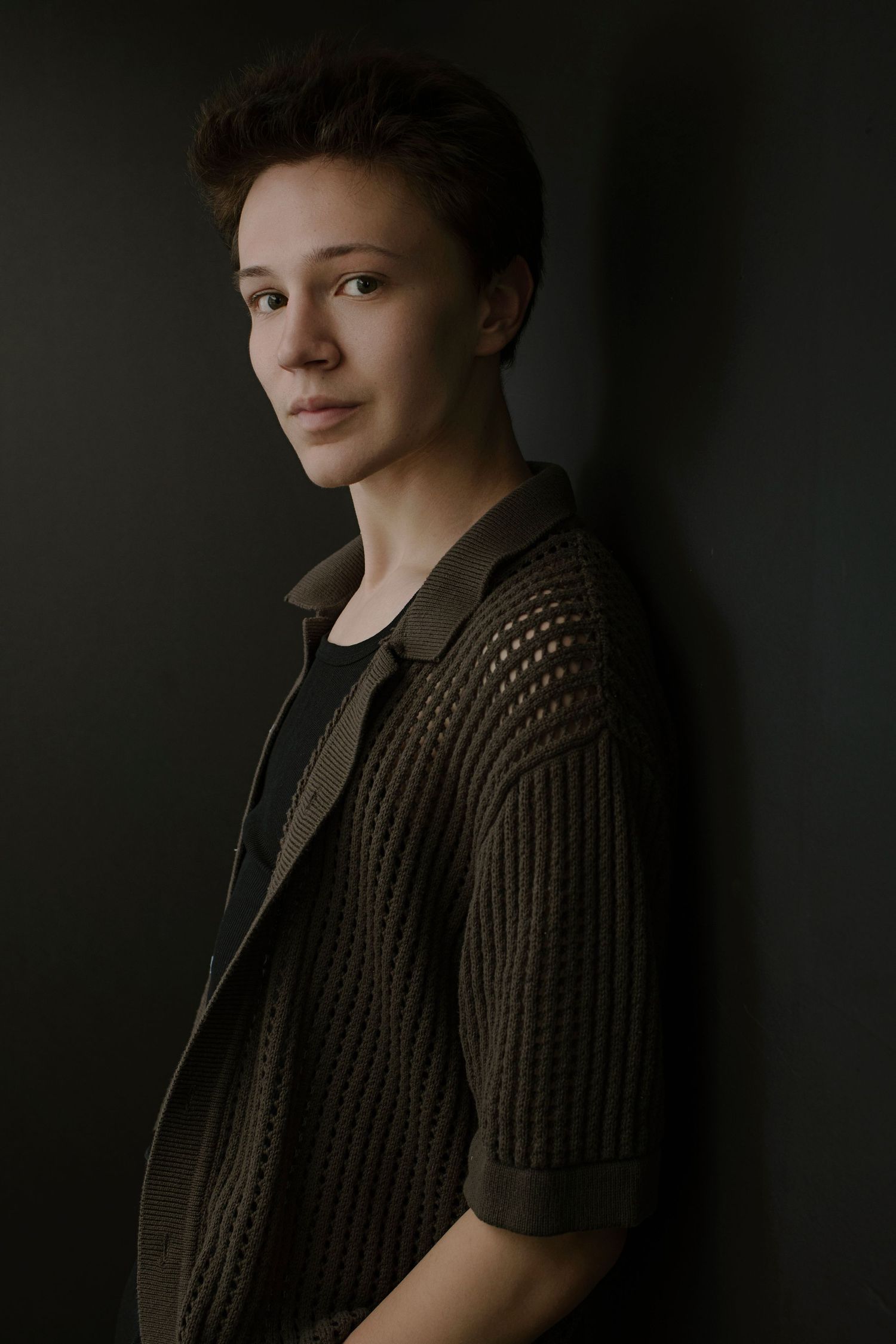 Young person with short dark hair in olive knit cardigan leans against dark wall looking at camera.