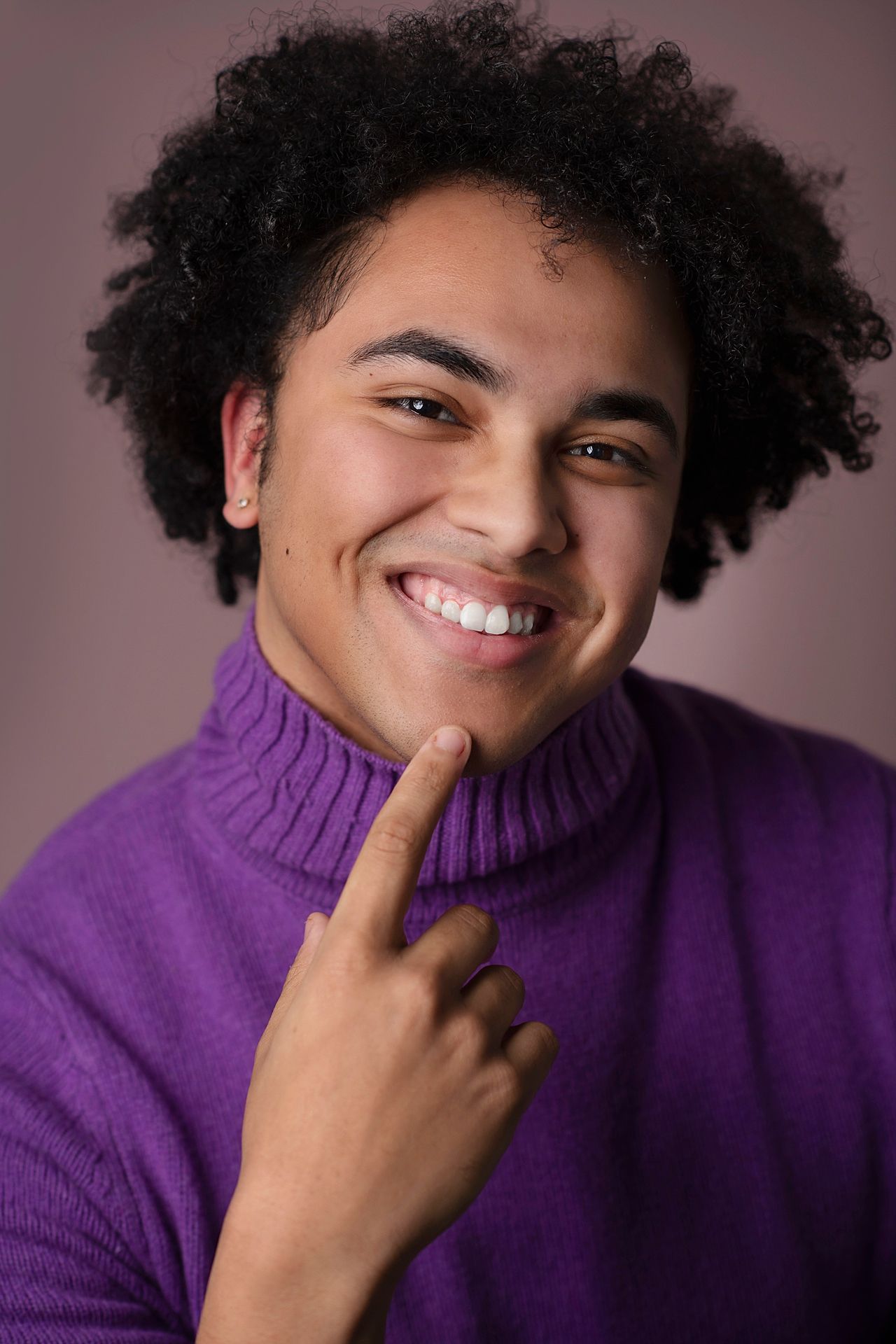 Cheerful young man with afro hair in a purple turtleneck pointing to his smile in a studio.