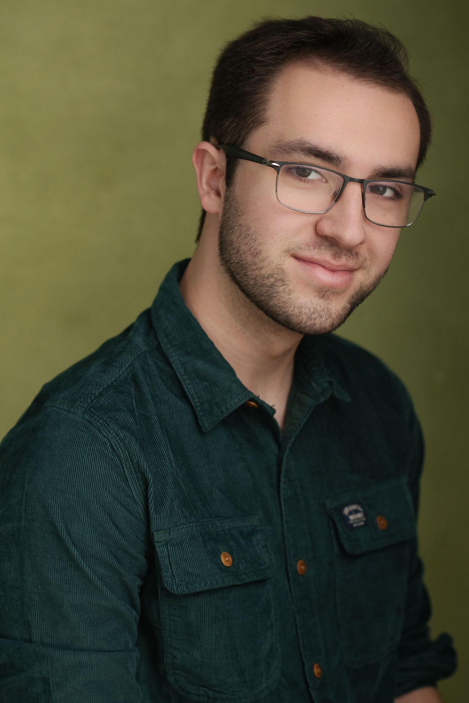 Young man with glasses and short beard smiling in green corduroy shirt against olive background.