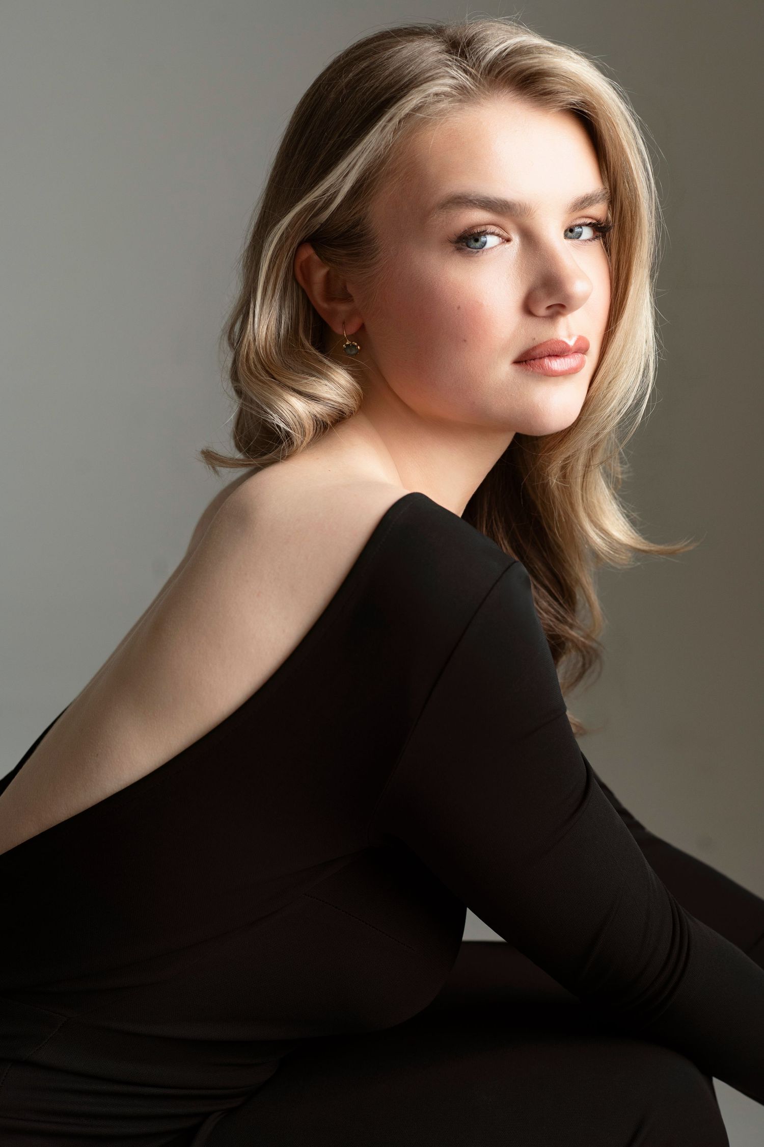 Young blonde woman in black off-shoulder top poses gracefully against gray background.