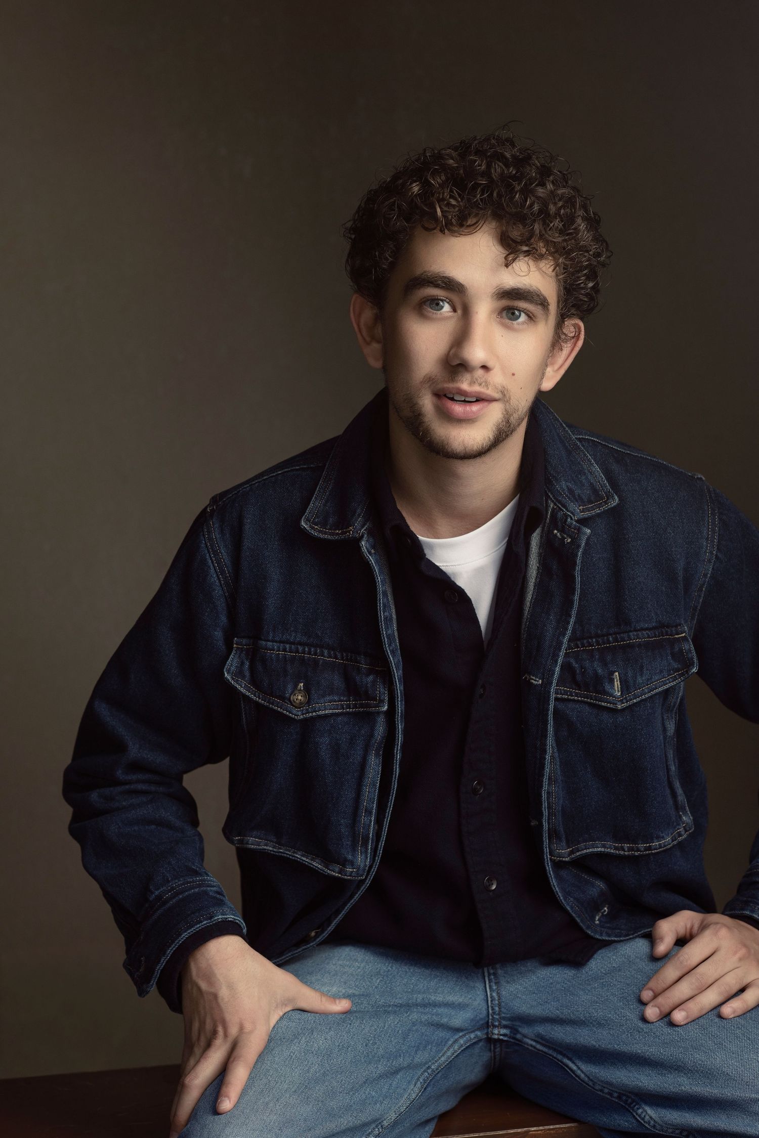 Young man with curly hair in denim jacket sits casually against olive background in studio portrait.