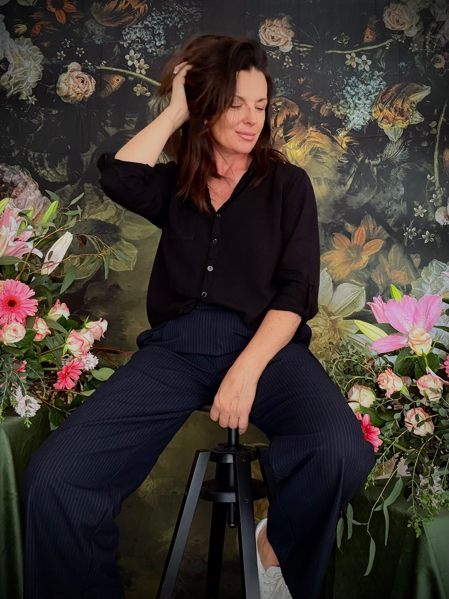 Brunette woman in black outfit sits on stool before floral dark botanical wallpaper backdrop.