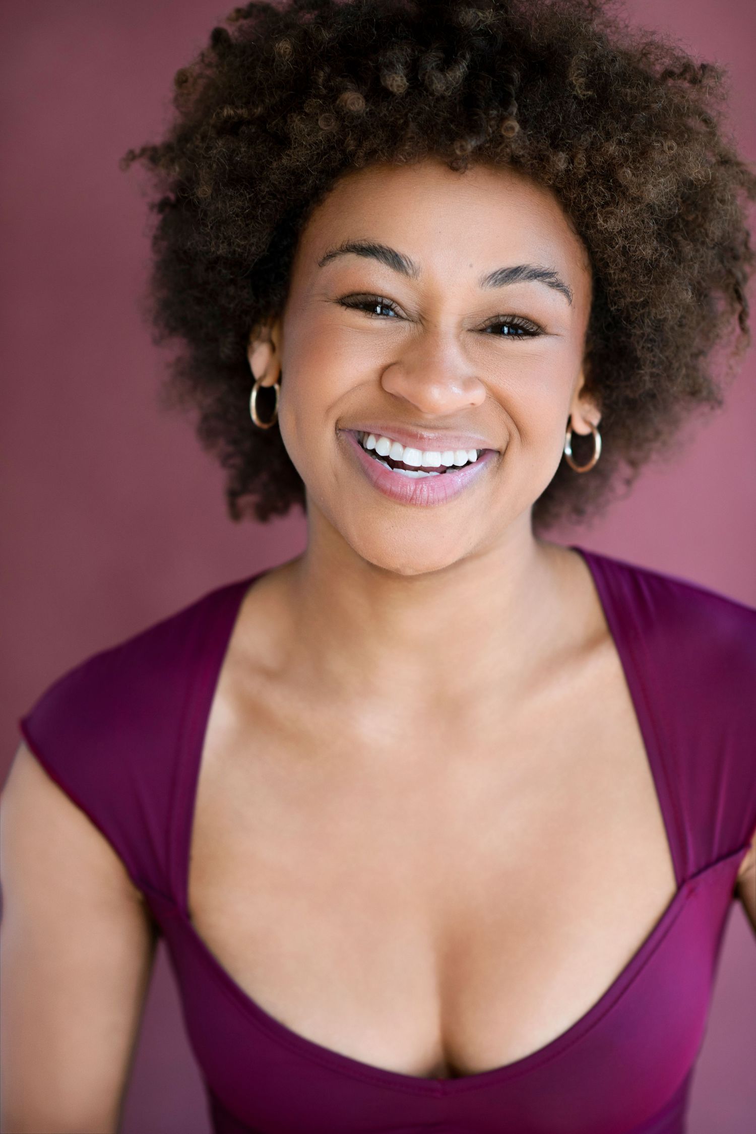 A young Black woman with a natural afro hairstyle smiles broadly, wearing a magenta top and gold hoop earrings.