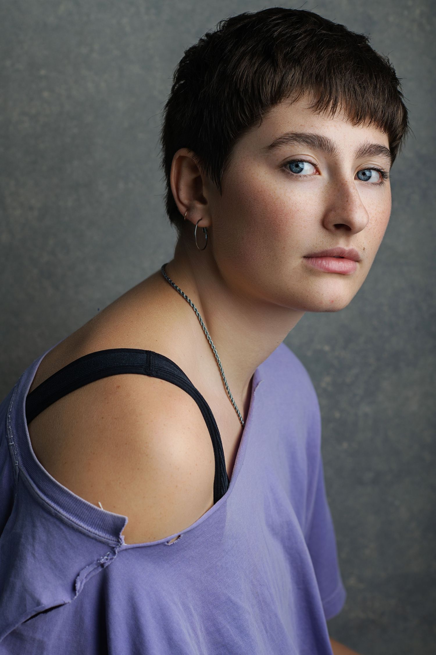 Young woman with short dark hair wearing a purple off-shoulder top and silver necklace poses against gray background.