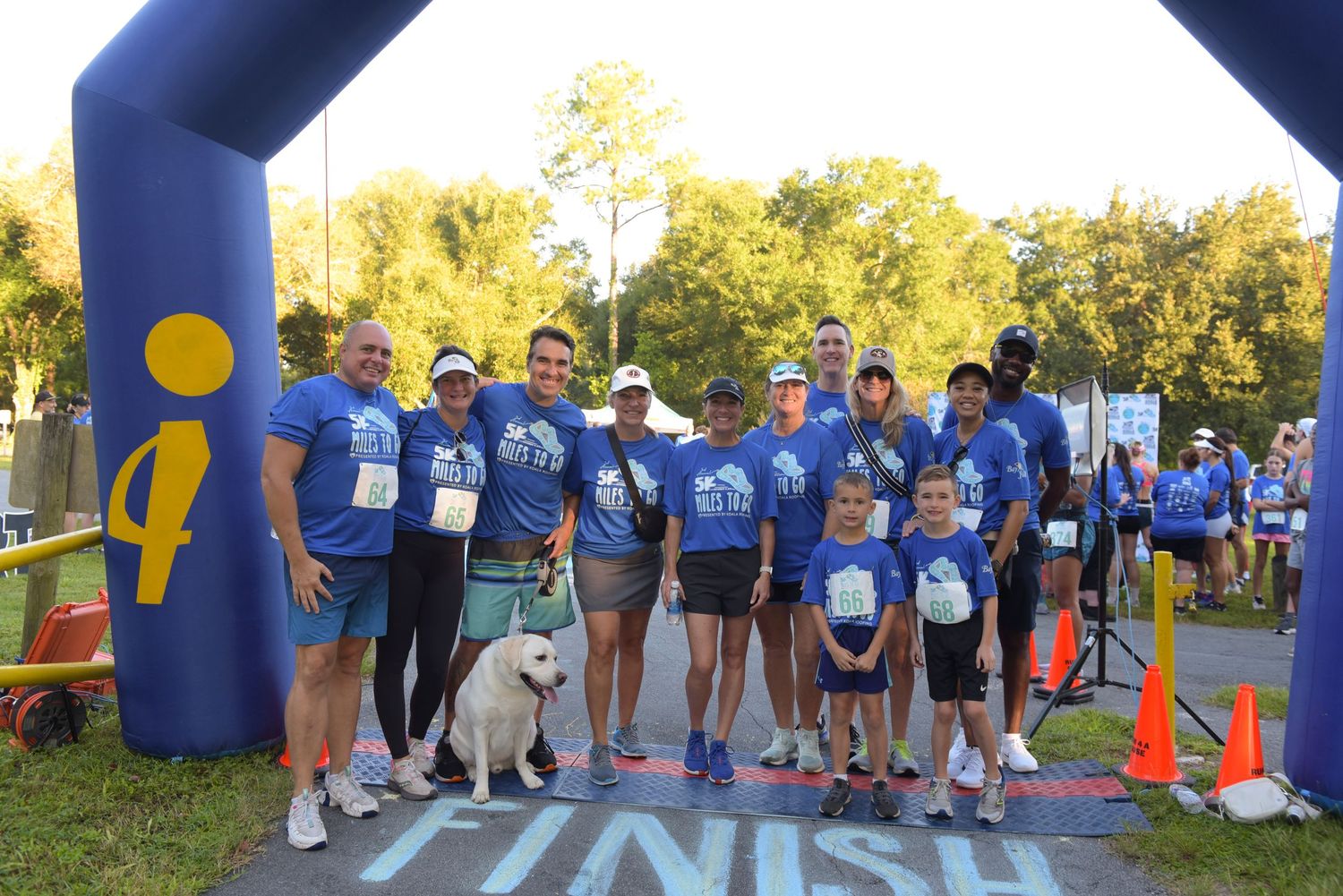 Group of people in blue t-shirts posing under a Finish line arch at a charity run event with a dog.