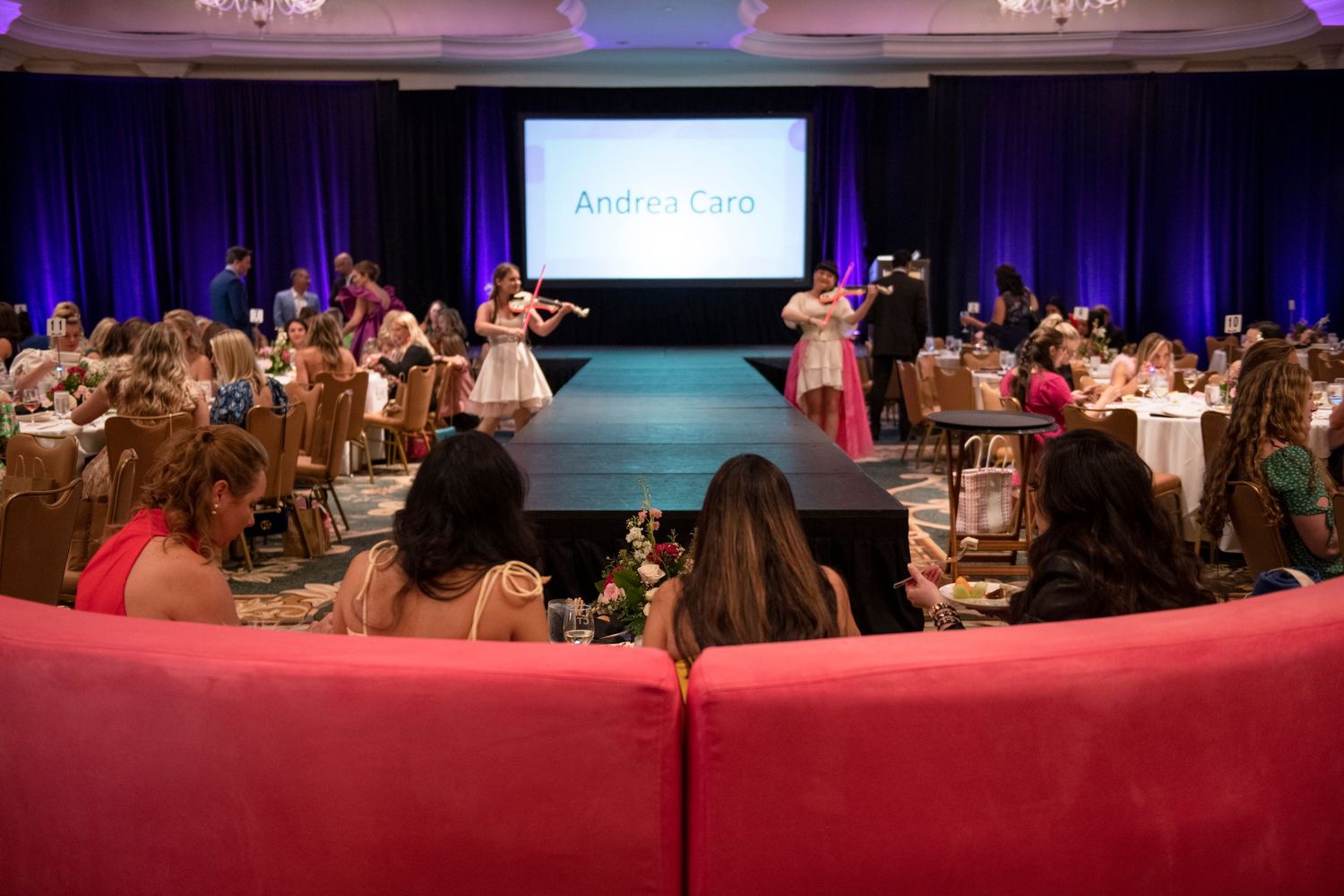 Elegant fashion show event in a ballroom with runway, violinists, and guests seated at decorated tables.