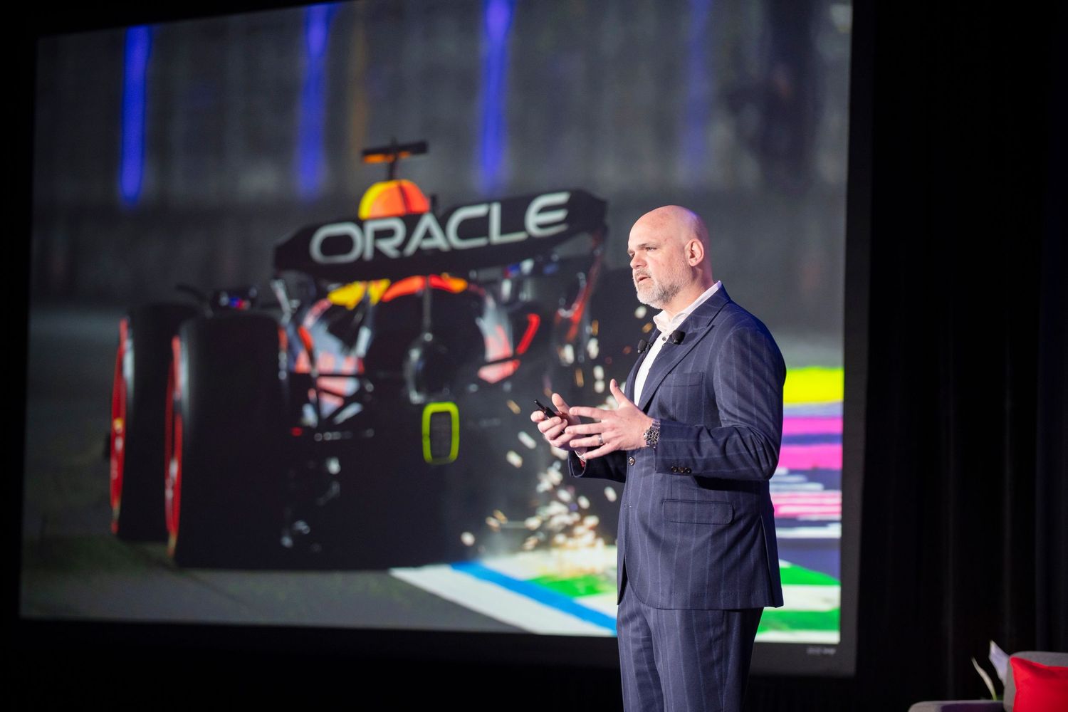 Executive presenting at Oracle Red Bull Racing F1 event, speaking on stage with race car projected on screen behind him.