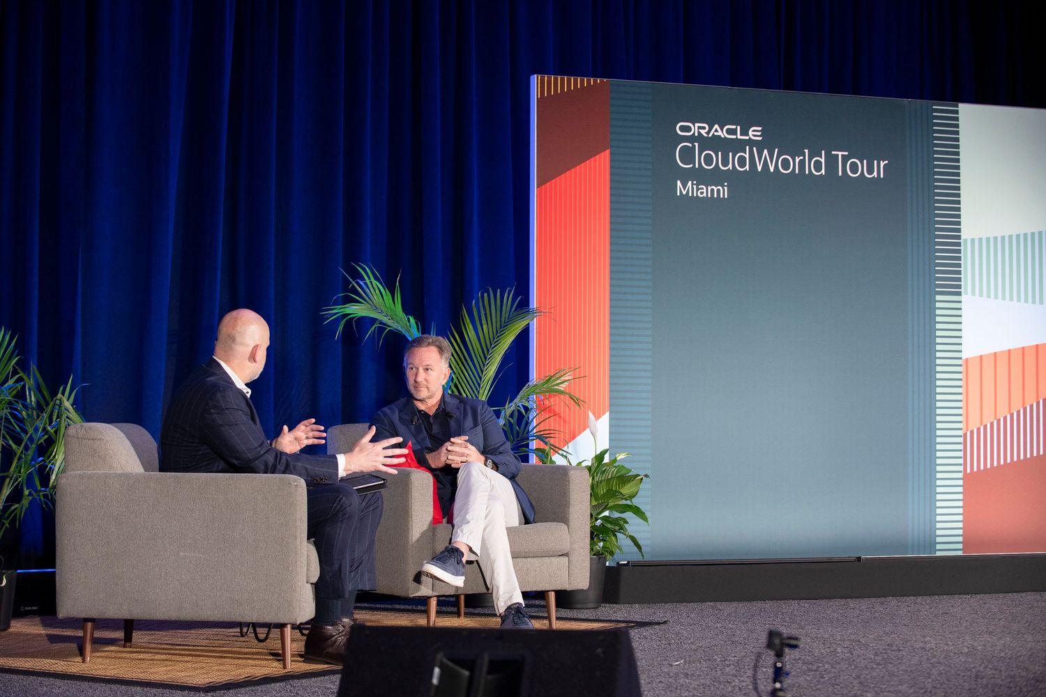 Two speakers on stage at Oracle CloudWorld Tour Miami event, seated in chairs against a branded backdrop.