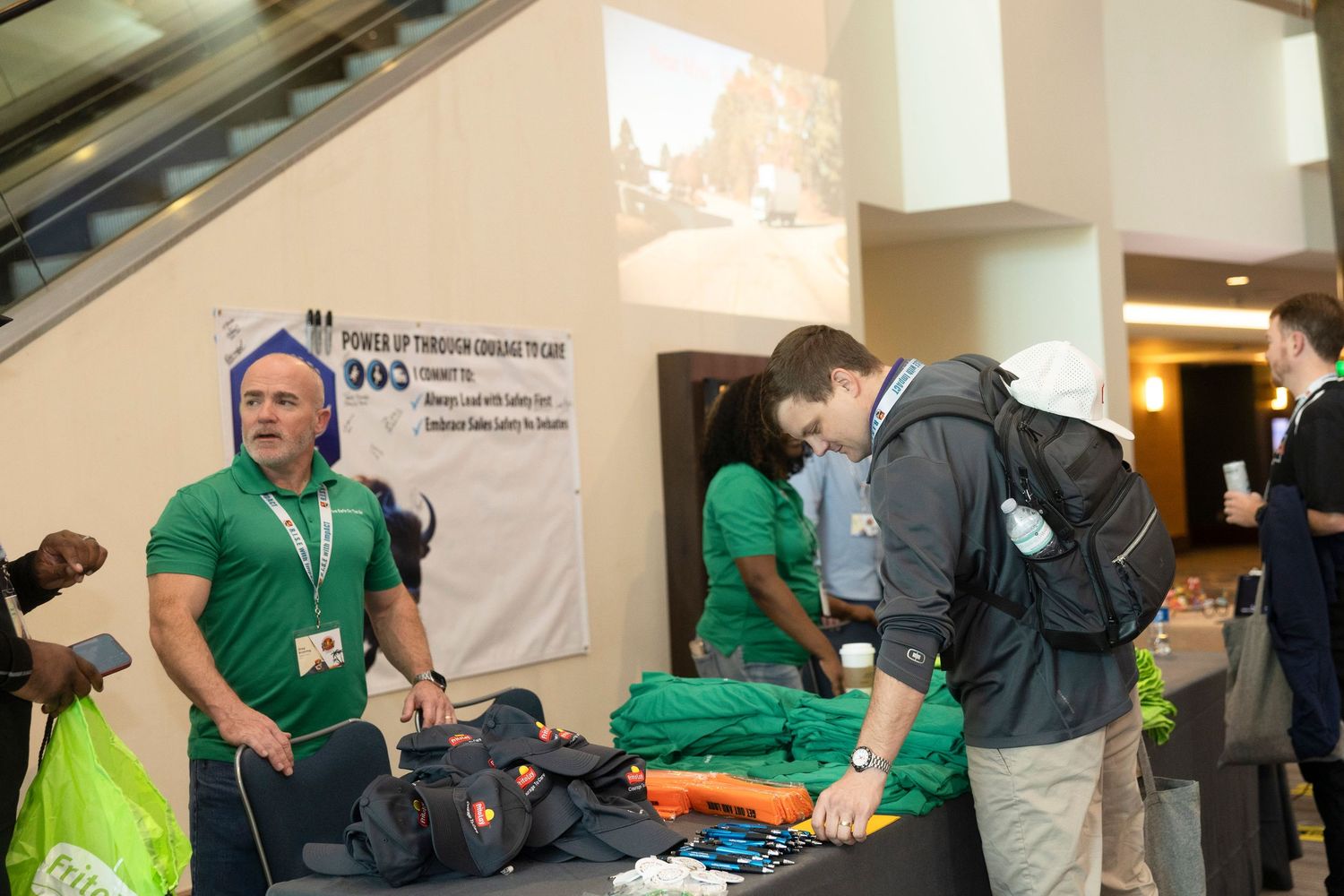 Volunteers in green shirts man a merchandise table at an indoor conference or charity event.