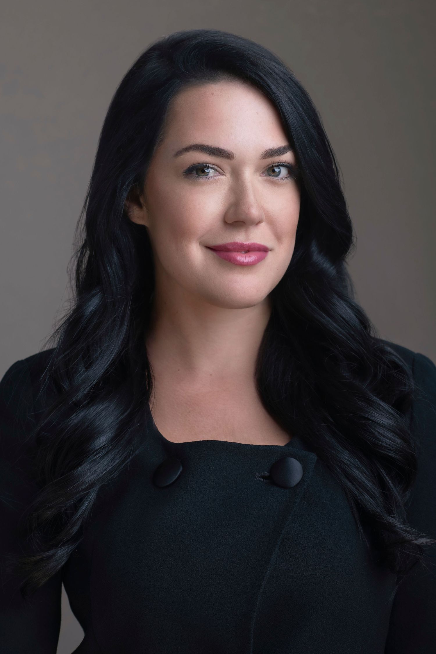 Professional headshot of a woman with long dark hair wearing a black blazer, smiling confidently against a neutral background.