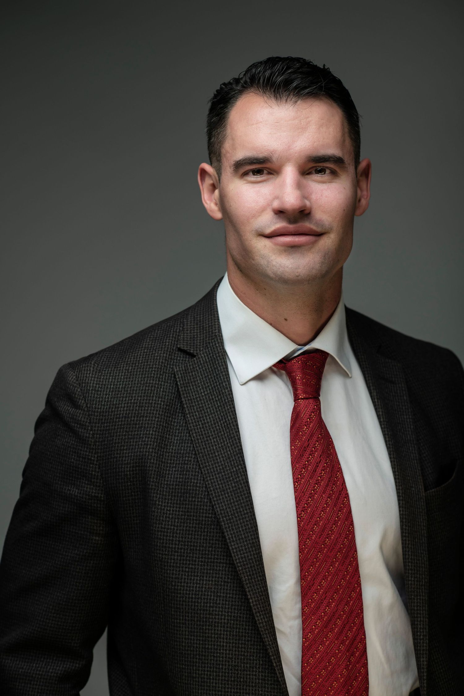 Professional man in dark suit and red tie posing for corporate headshot against gray background.