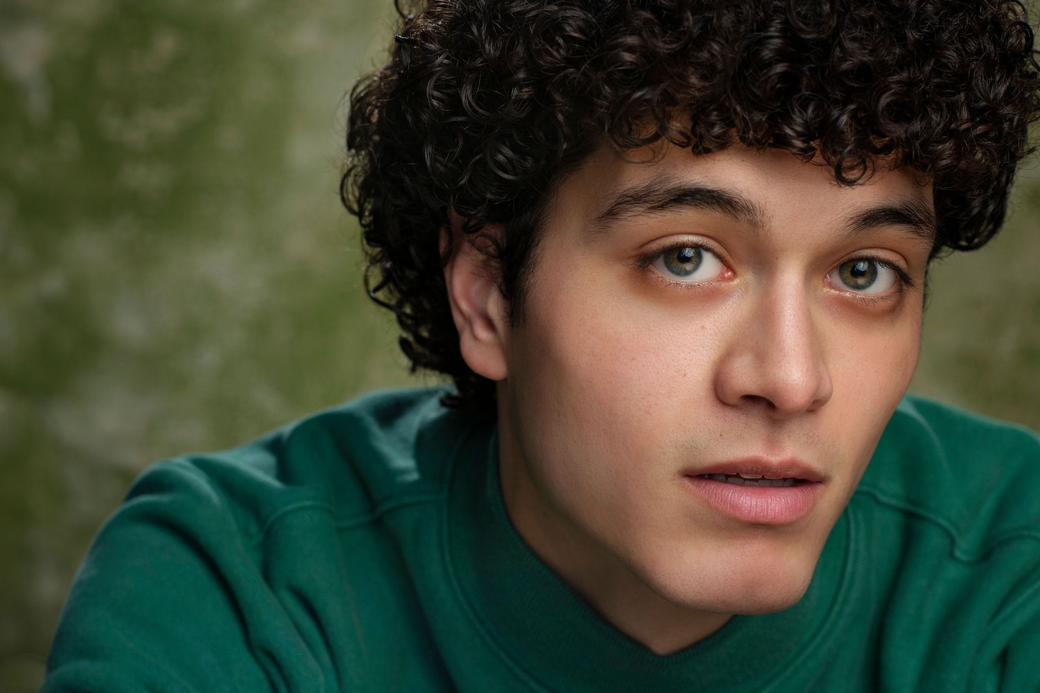 Close-up portrait of a young man with curly dark hair wearing a green sweatshirt against a textured background.
