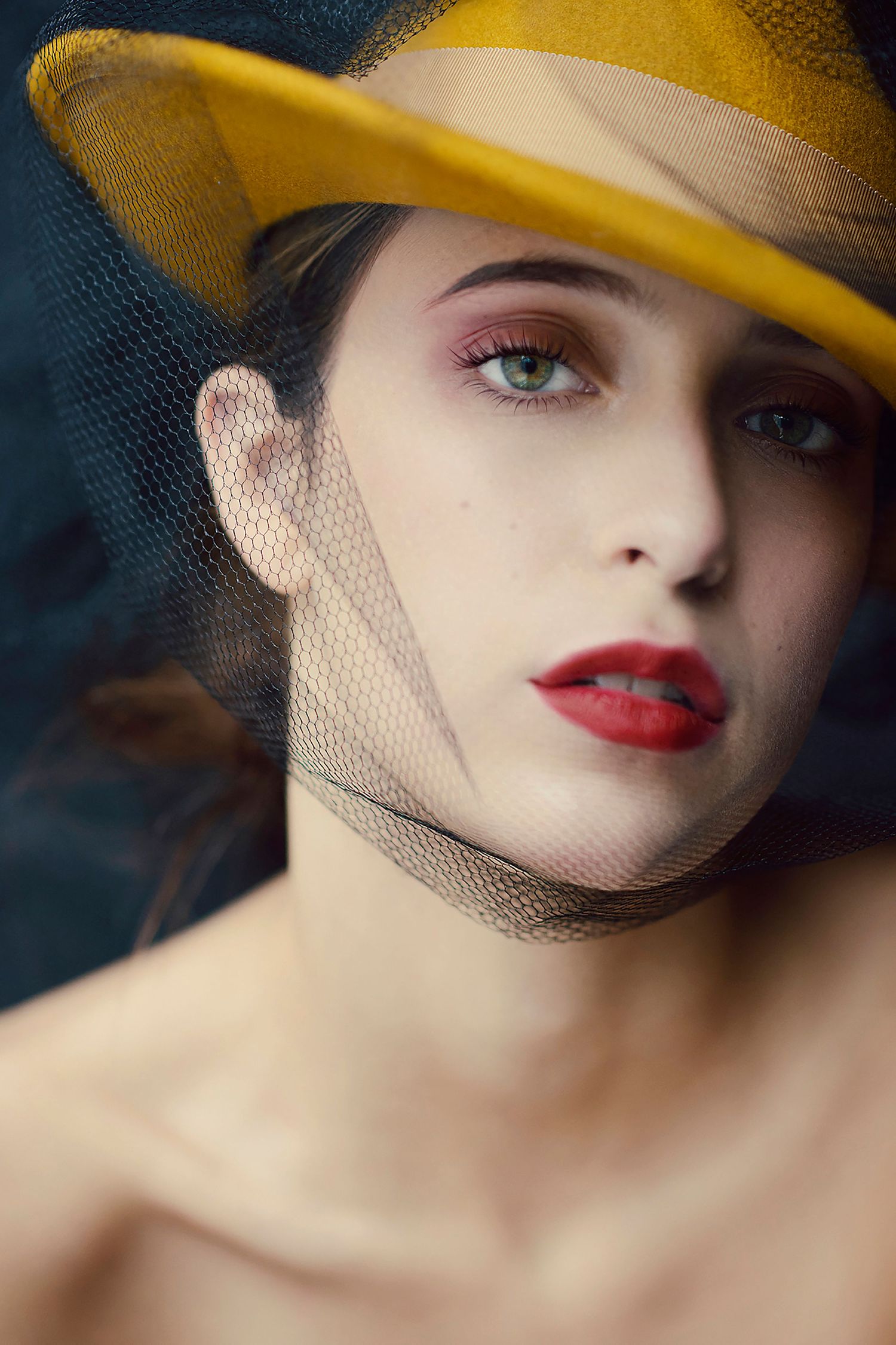 Portrait of a young woman wearing a yellow hat with black mesh veil and red lipstick.