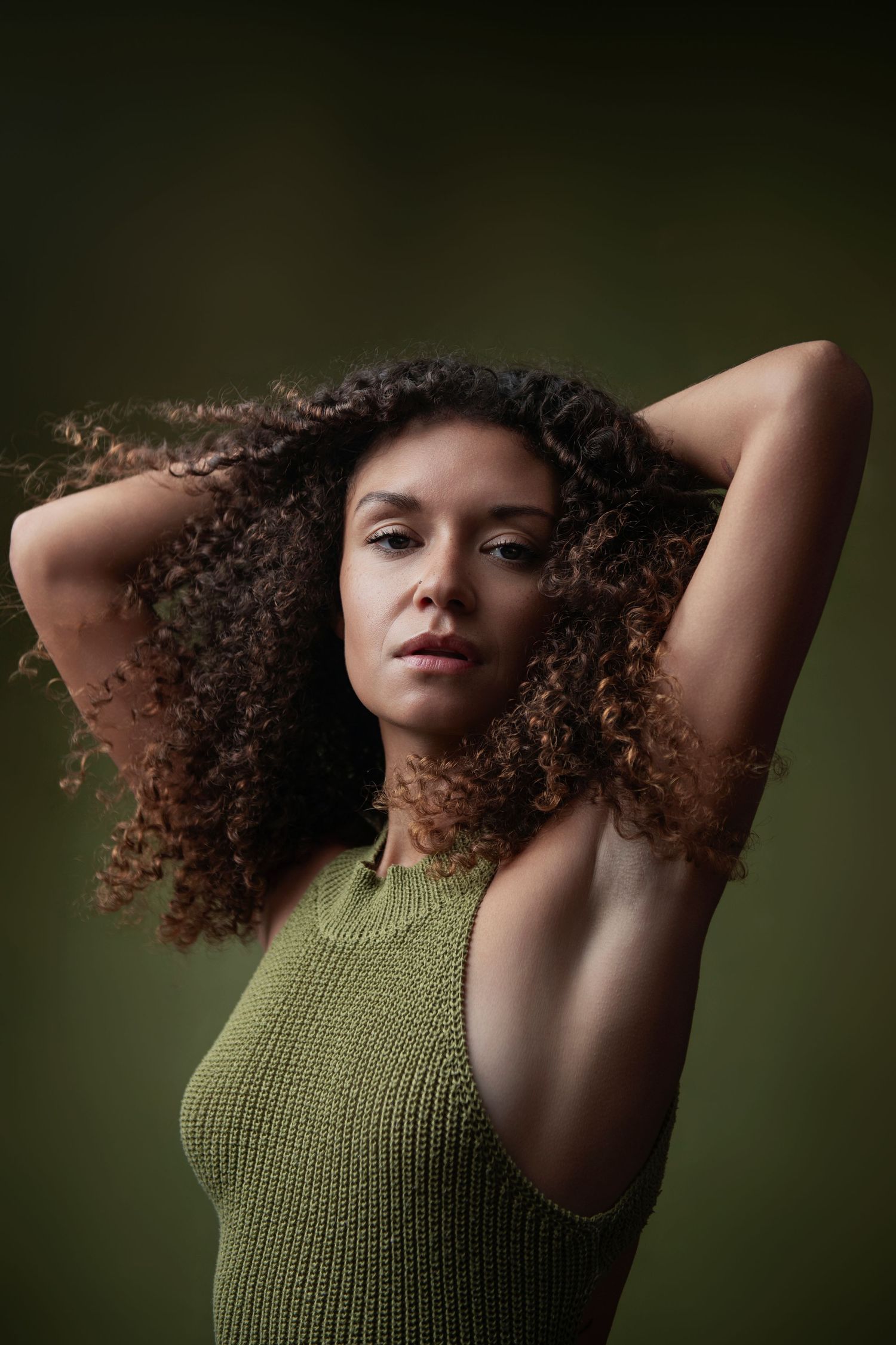 Woman with curly hair in green knit top poses with hands behind head against dark green background.