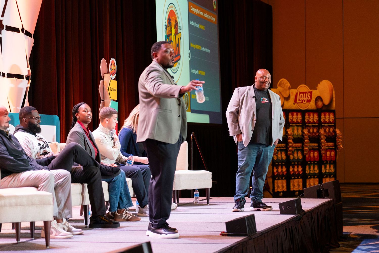 Two men present on a conference stage with seated audience and colorful display backdrop.