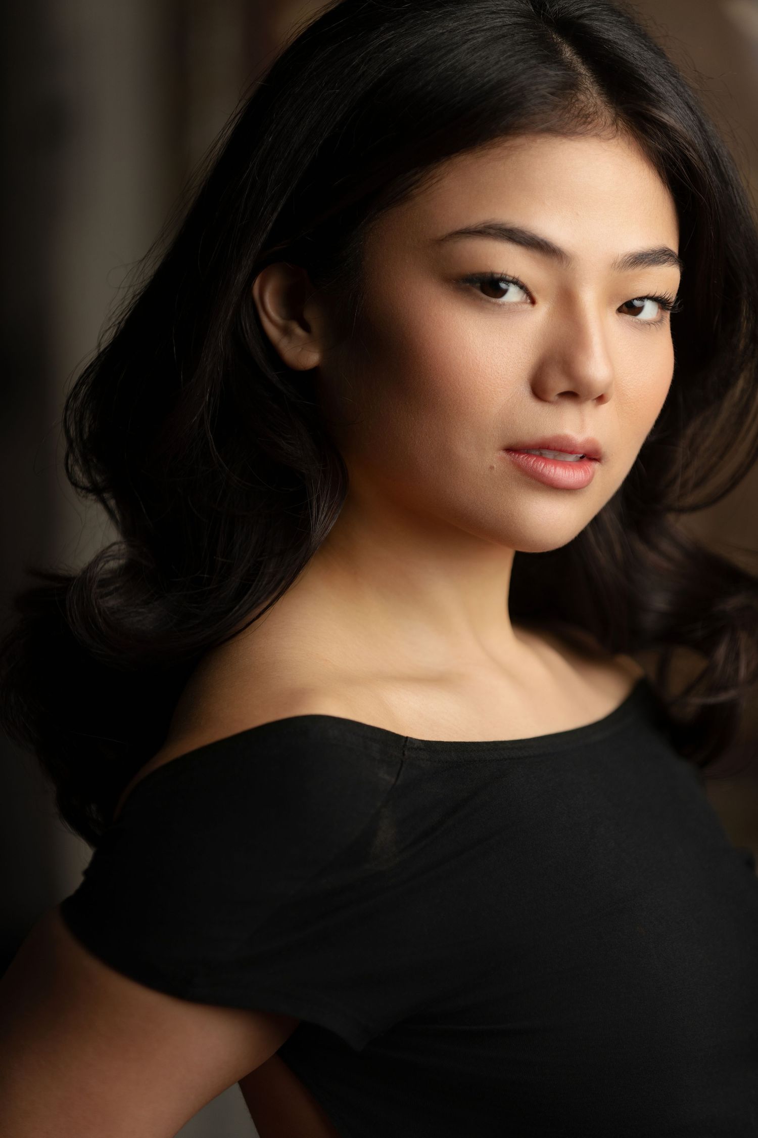 Young Asian woman with long dark hair wearing an off-shoulder black top, posing for a professional portrait photo.