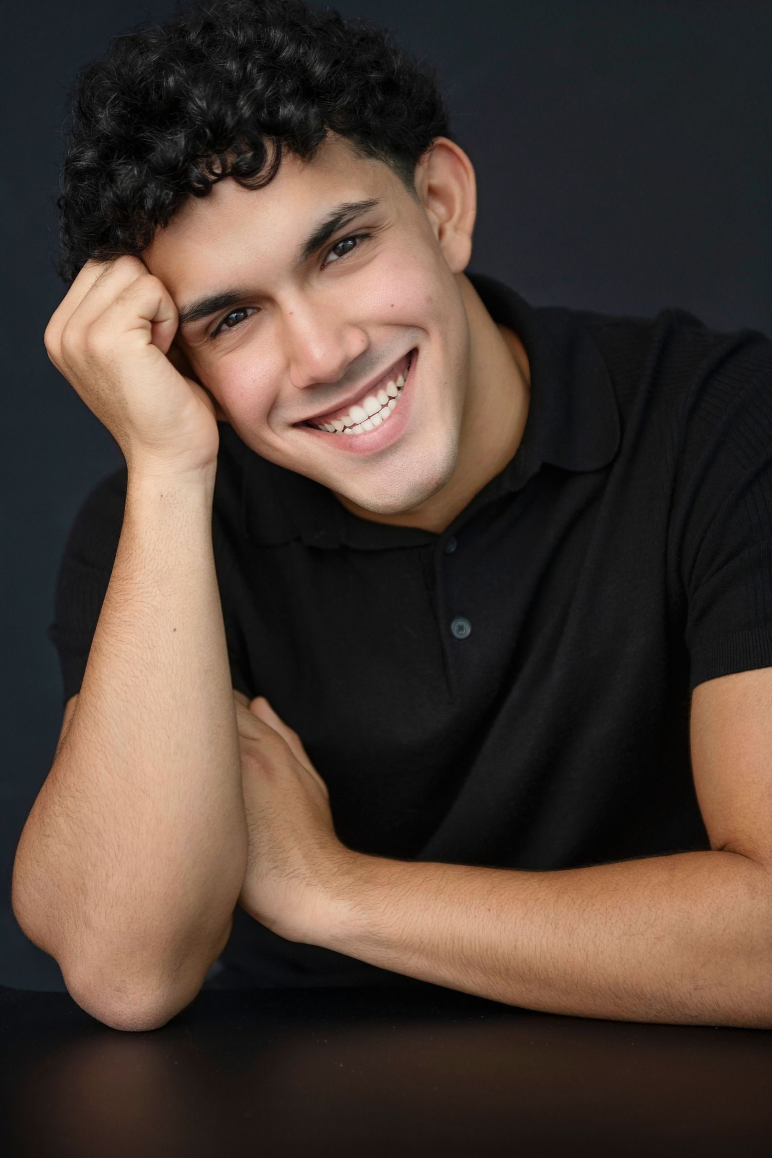 Young Hispanic man with curly hair smiling warmly while resting head on hand against dark background.