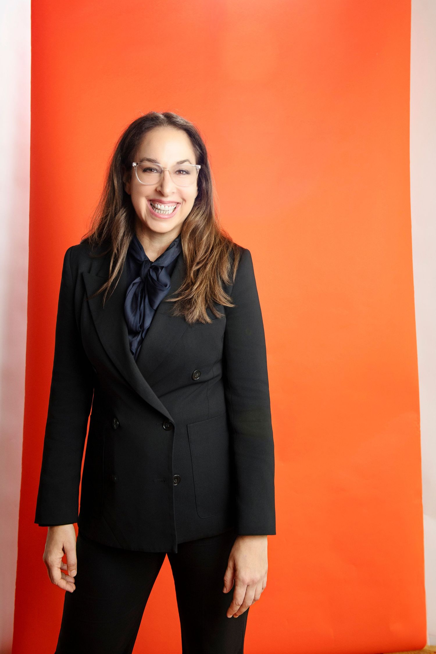 Smiling woman in black blazer and navy bow tie, wearing glasses, posing against a bright orange background.