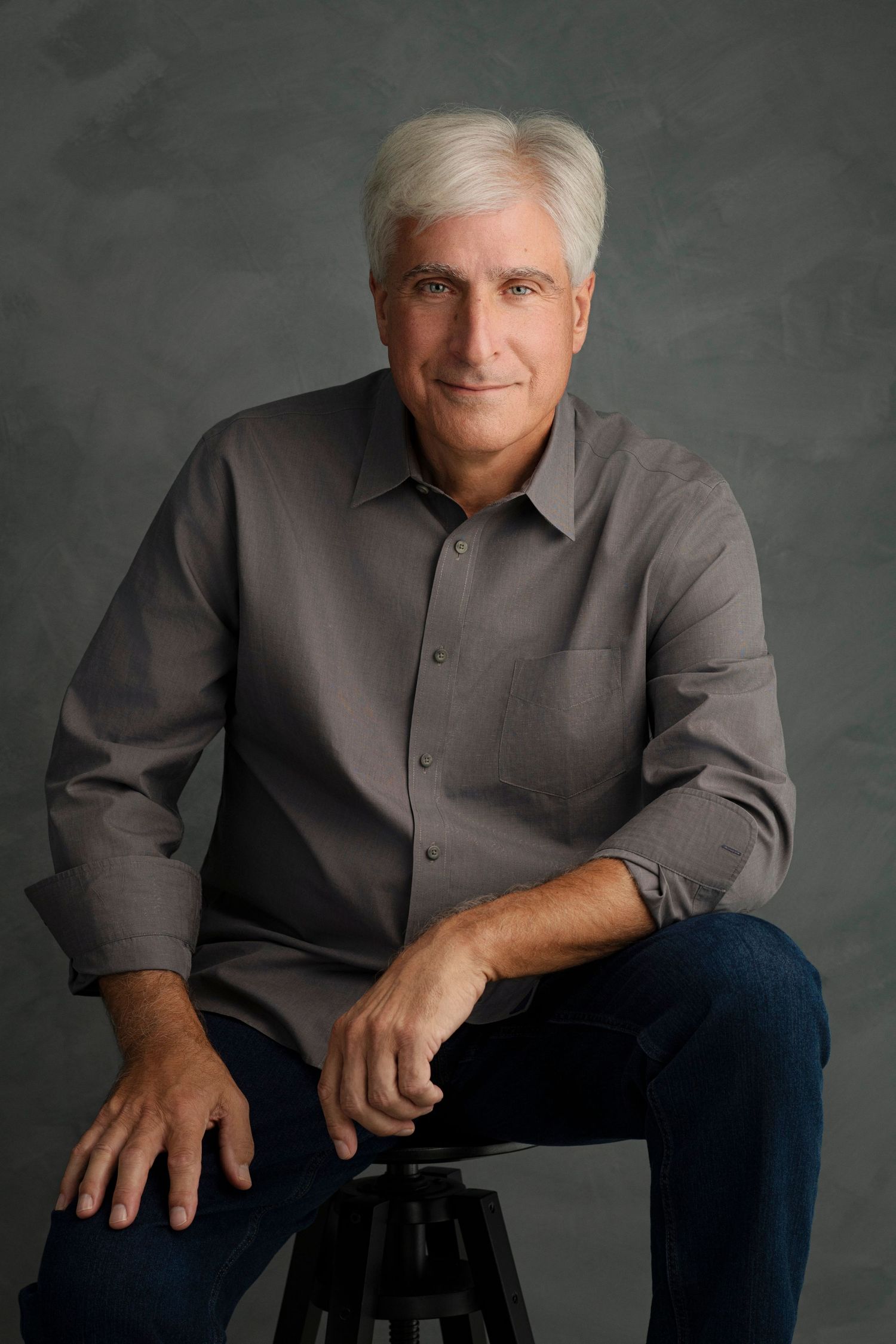 Middle-aged man with white hair sitting on stool, wearing gray shirt and jeans, smiling confidently in studio portrait.