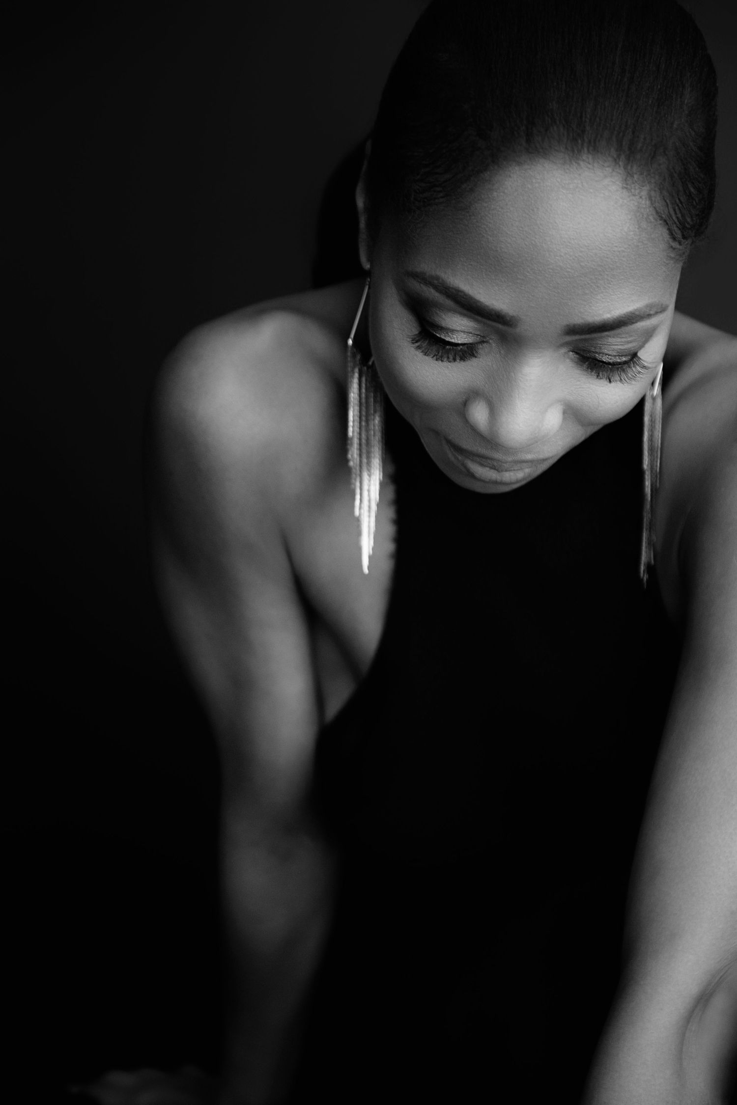 Black and white portrait of a woman in a black dress with long earrings, looking down against a dark background.