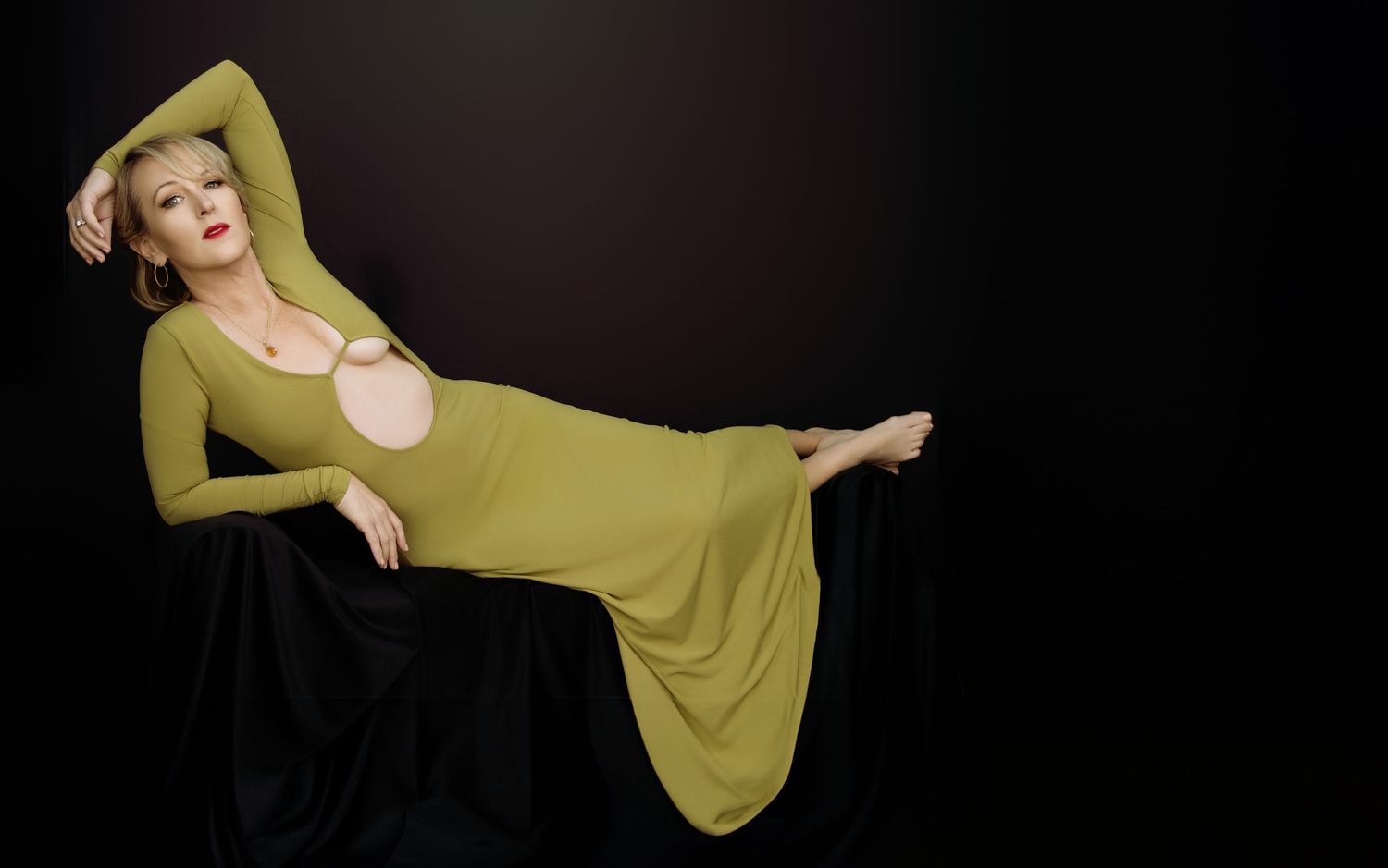Woman in olive green long-sleeve cutout dress poses elegantly against dark background.