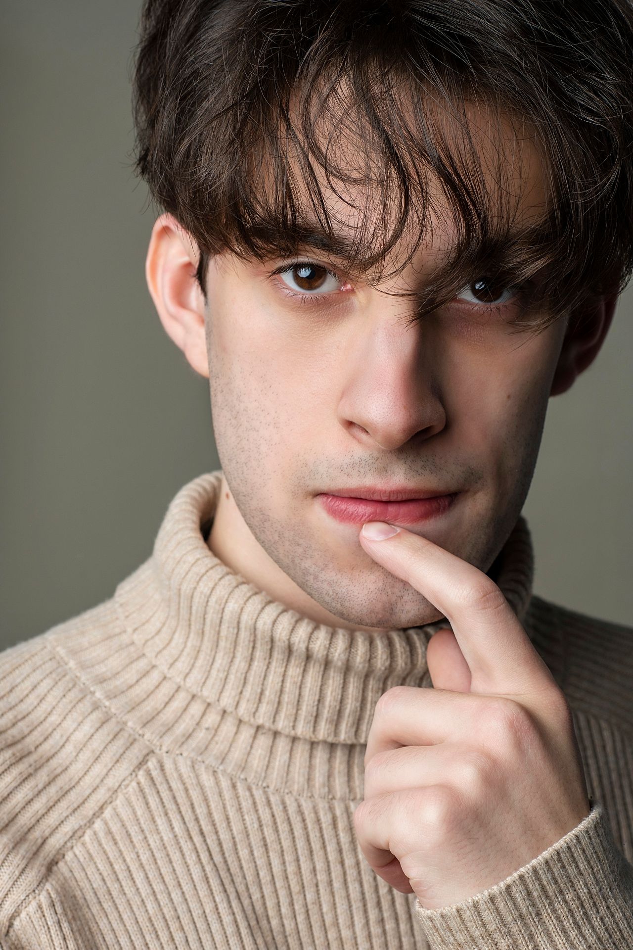 Intense young man with dark hair in a beige turtleneck touches his lip in a studio portrait.