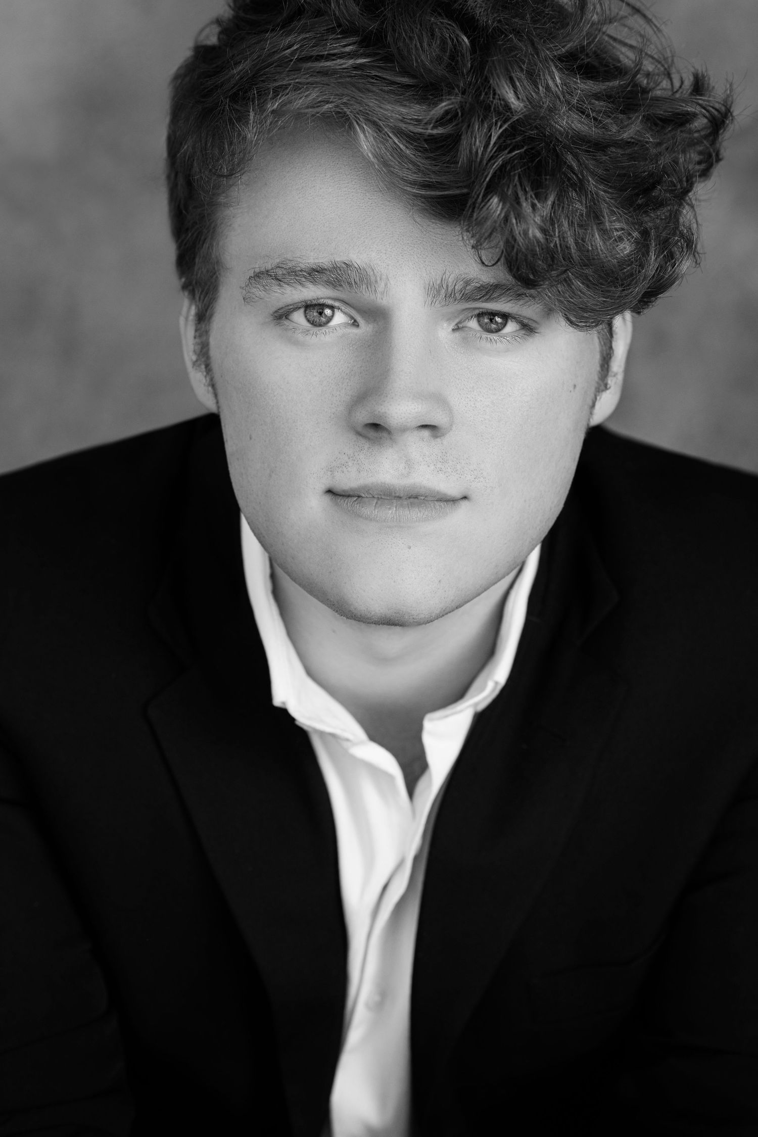 Black and white headshot of a young man with curly hair wearing a black blazer and white shirt.