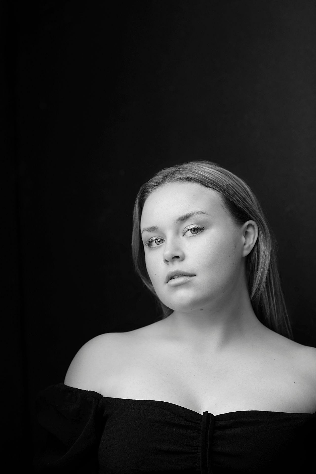Black and white portrait of a young blonde woman in an off-shoulder top against dark background.