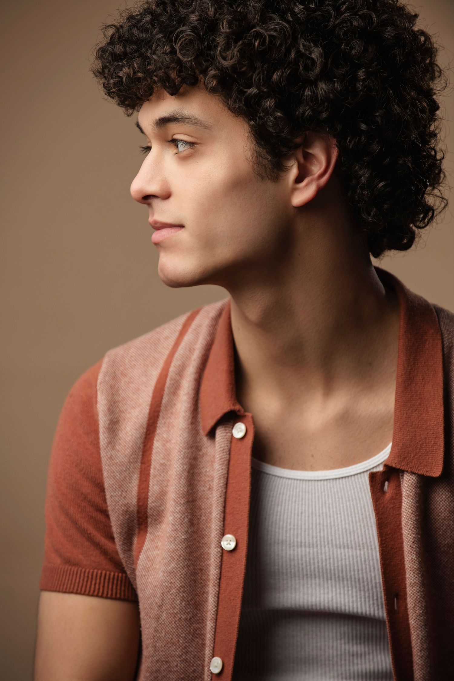 Side profile portrait of a young man with curly dark hair wearing a brown button-up short-sleeve shirt.