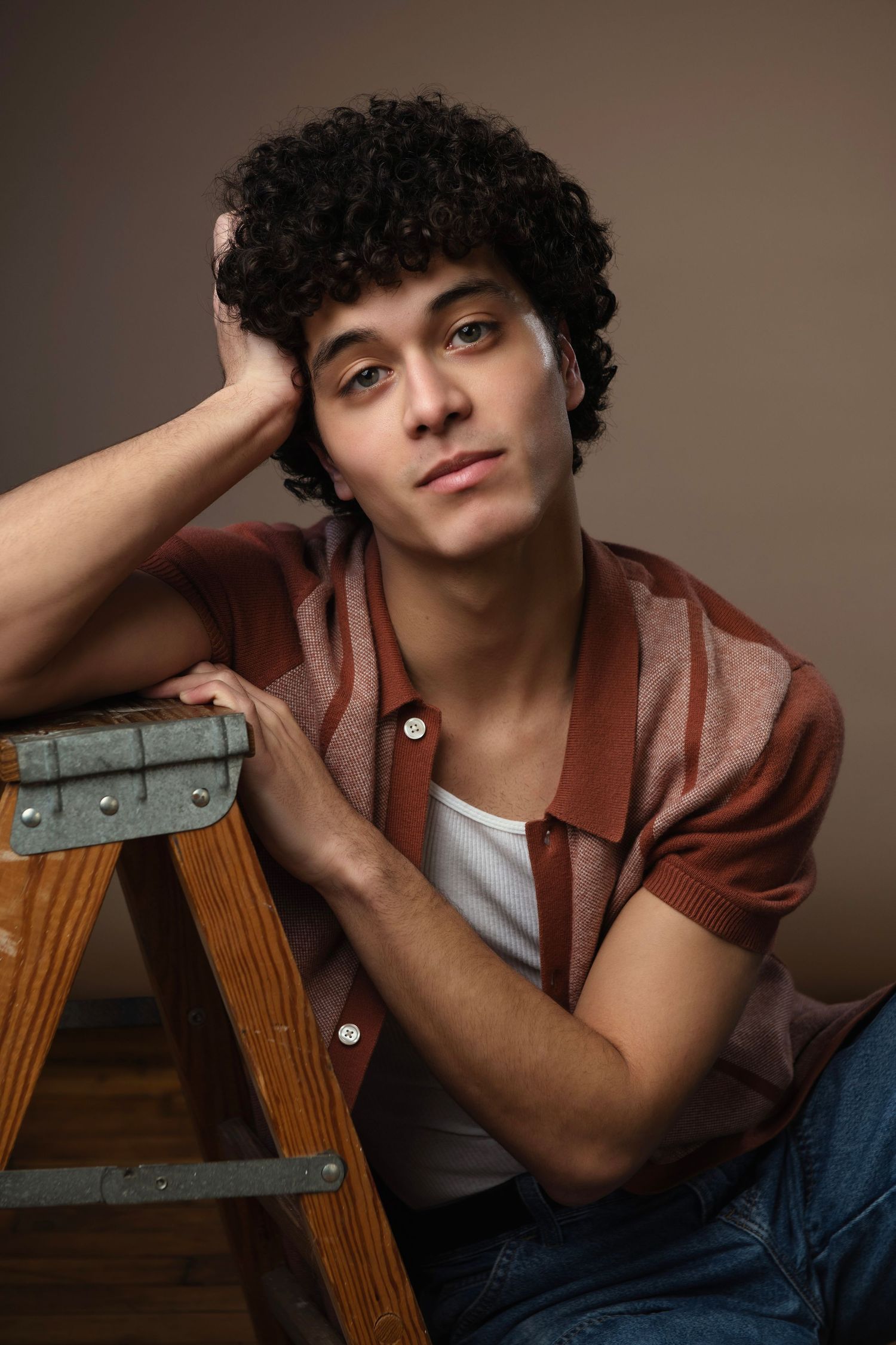 Young man with curly dark hair leaning on a wooden ladder, wearing a brown striped polo shirt and jeans.