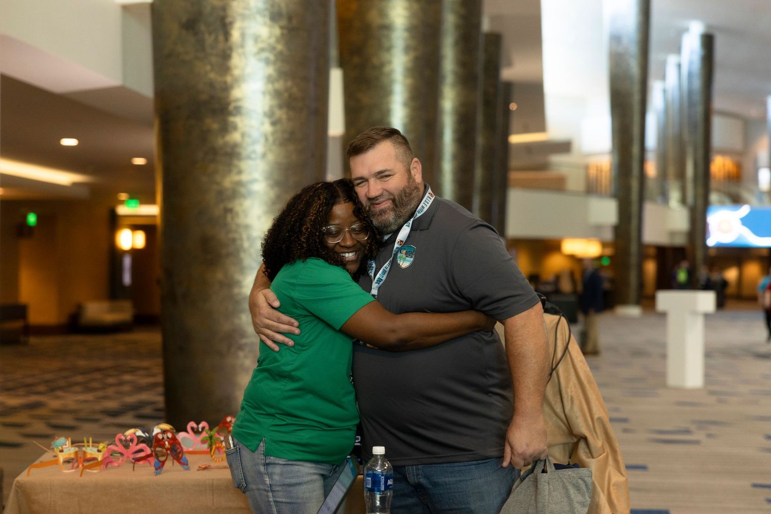 Two people share a warm hug in a modern lobby, both wearing green shirts, with colorful items on a table nearby.