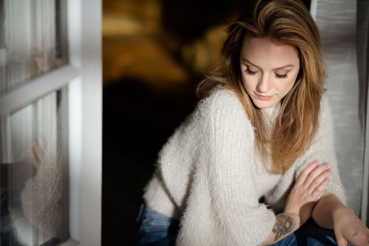 Woman with long wavy auburn hair in a fuzzy cream sweater and jeans sits by a window, gazing down with a wrist tattoo visible.