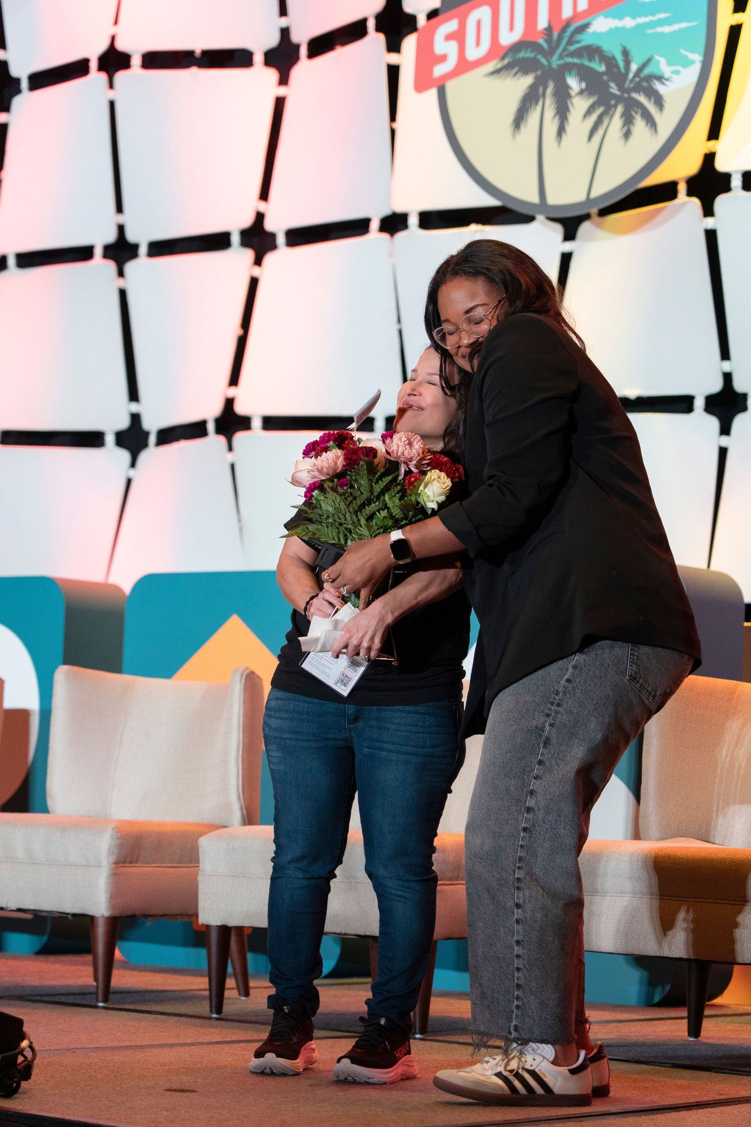 Two women share a warm hug on stage at a South by Southwest event, one holding a bouquet of flowers.
