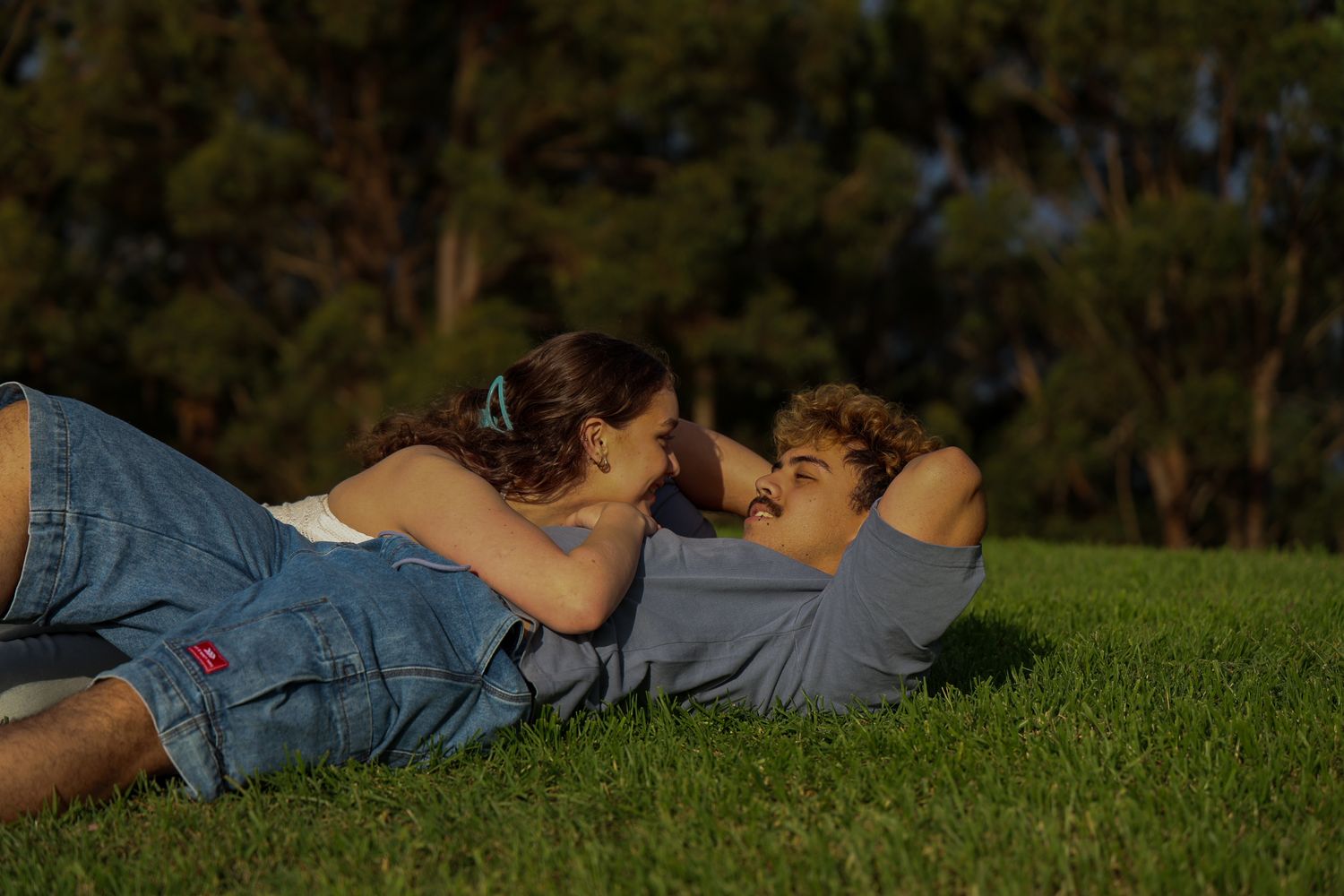 A young couple laying on green grass together at sunset, looking into each others eyes