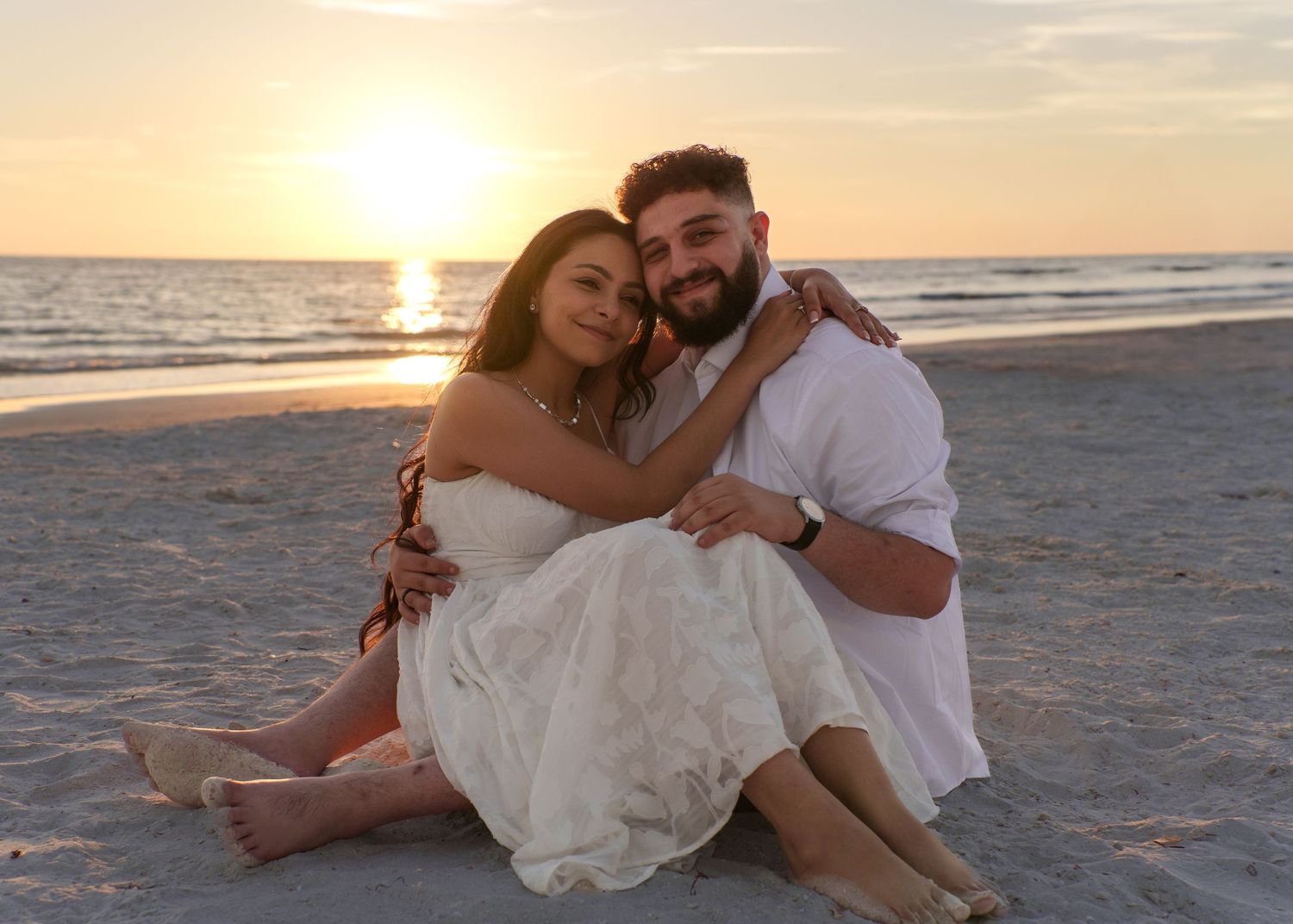 Romantic couple sitting on beach at sunset in casual attire.