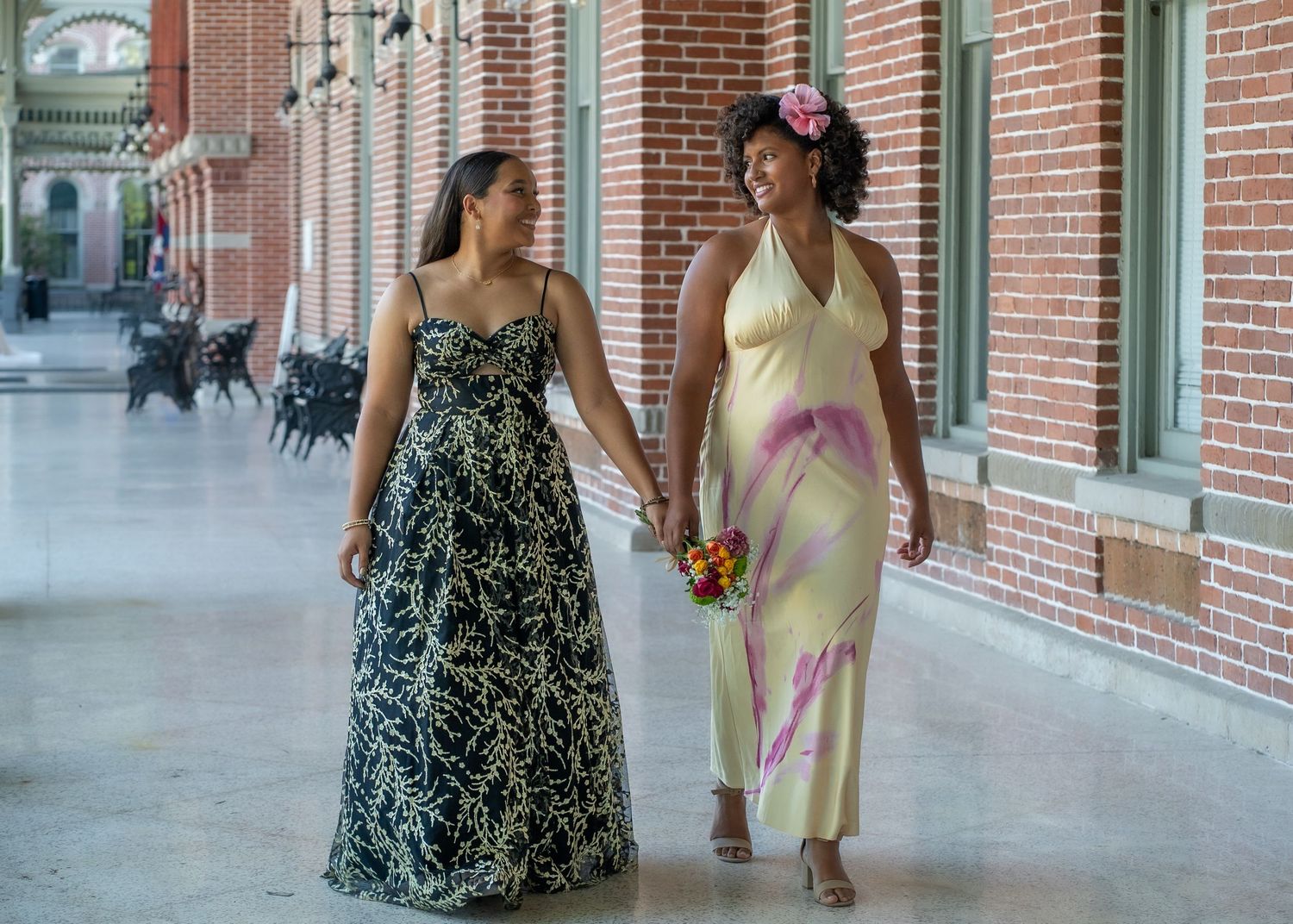 Two people walk together along a covered walkway, one wearing a yellow dress and one in black and white.