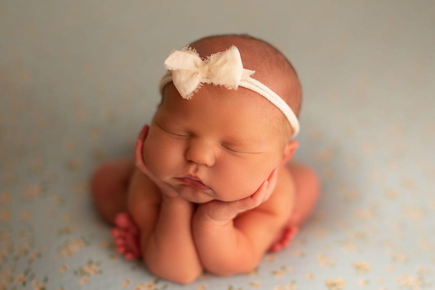Sleeping newborn baby girl wearing a white bow headband, posed with hands under chin on a soft blue backdrop.