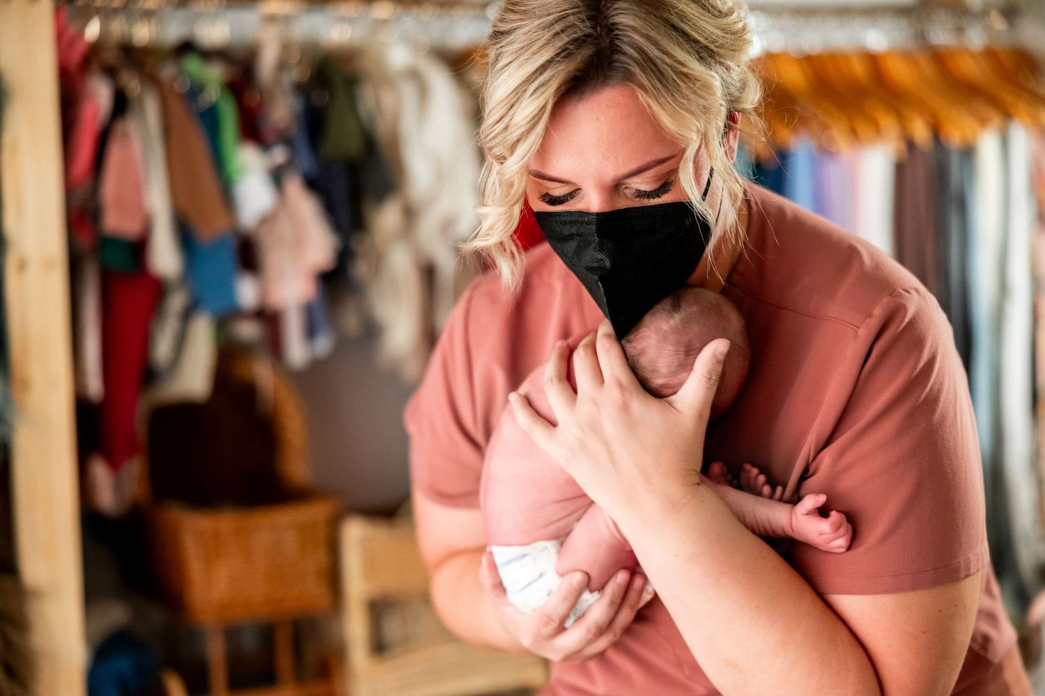 Woman wearing black face mask tenderly holds newborn baby in a rustic room with hanging clothes.