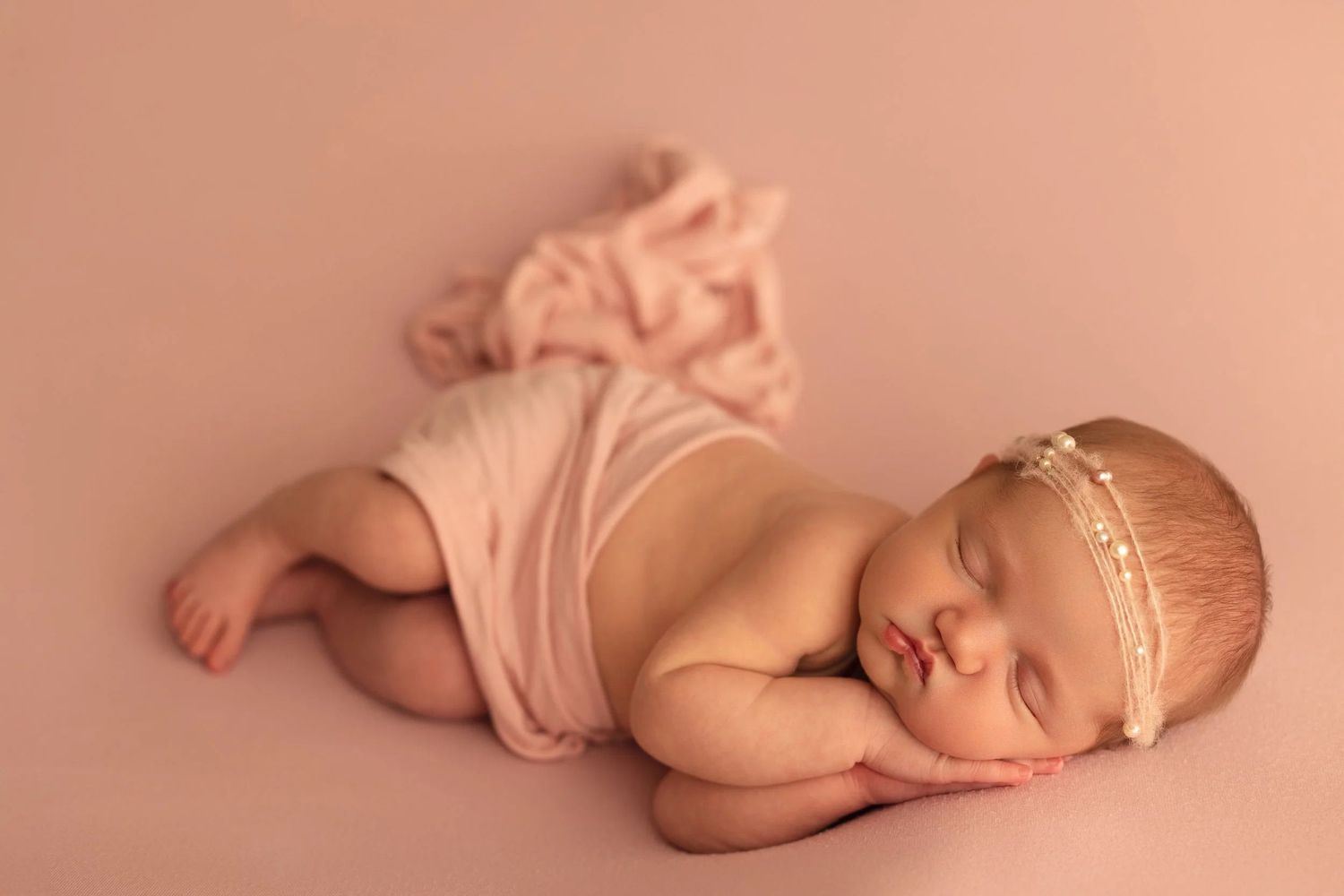 Peaceful sleeping newborn baby girl in pink wrap and gold headband on matching blush pink backdrop. Newborn Photography Texarkana TX