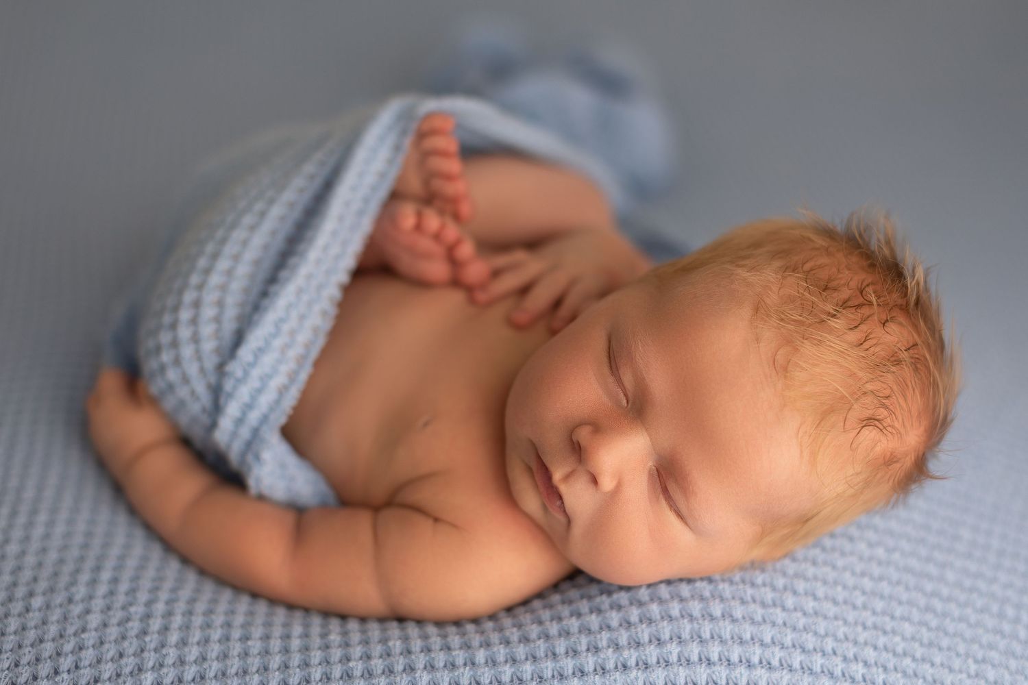 Smiling newborn baby boy wrapped in a soft blue blanket, peacefully sleeping on a blue knit surface.