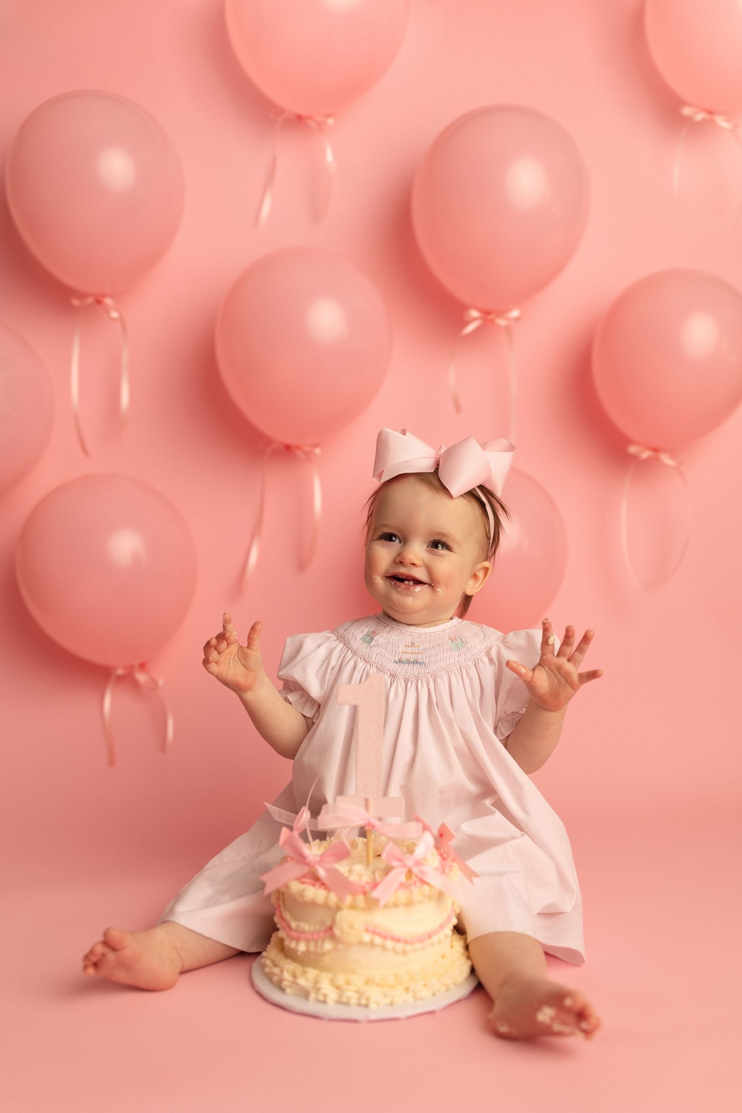 Smiling baby girl in white dress celebrates first birthday with pink cake and balloon backdrop. Milestone Photography Texarkana TX