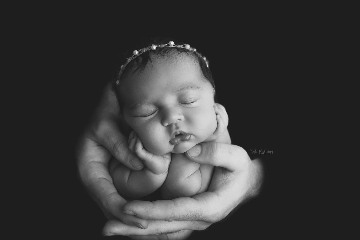 Black and white portrait of sleeping newborn baby cradled gently in parent's hands with delicate floral headband. Newborn Photographer