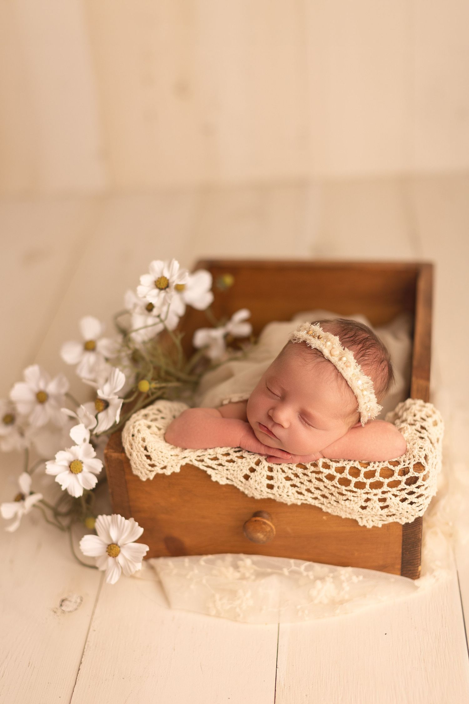 Sleeping newborn baby in wooden drawer with lace blanket and white flowers, wearing cream headband.