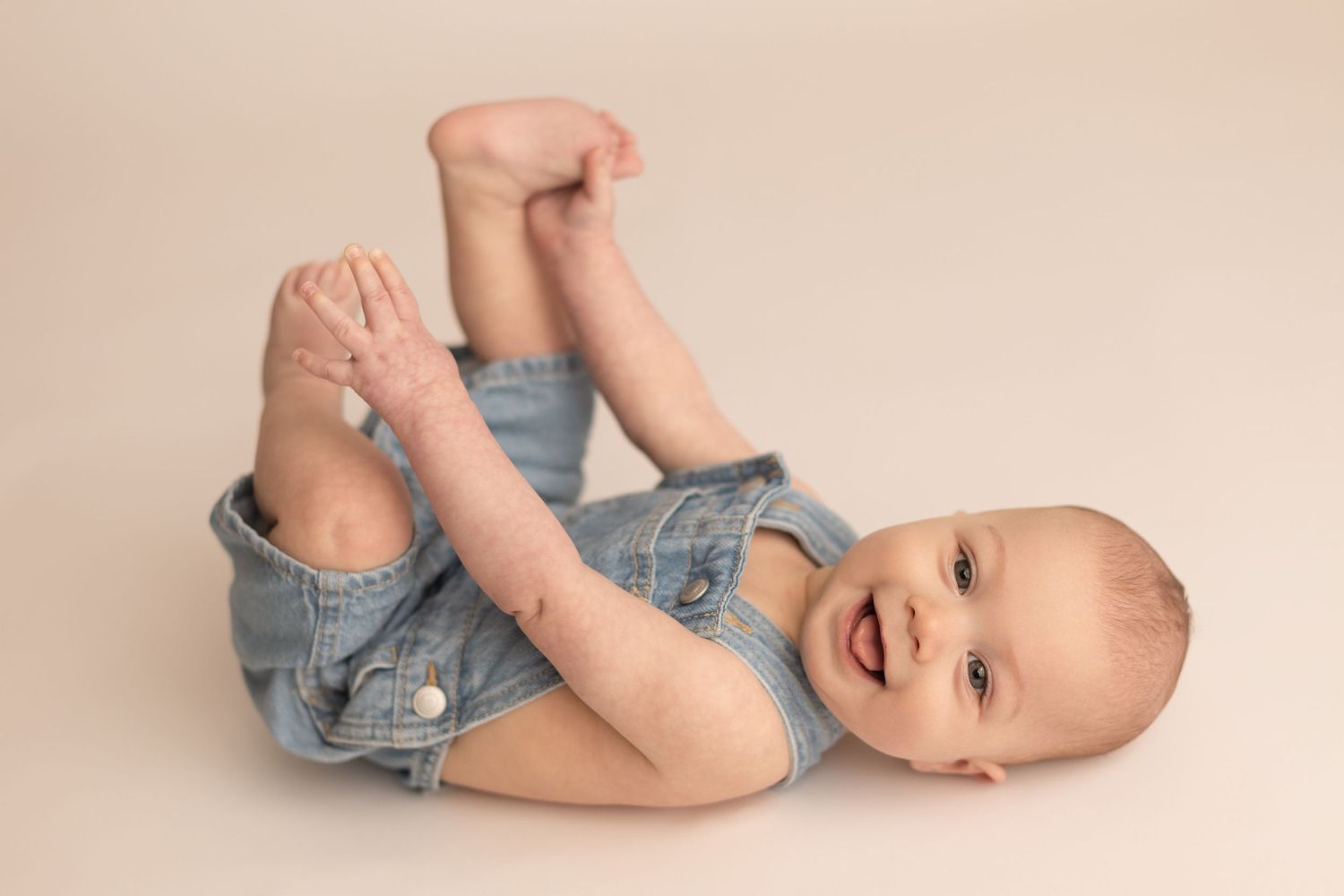 Happy baby in denim romper lies on back laughing while holding feet up in adorable milestone photo. Milestone Photography Texarkana TX