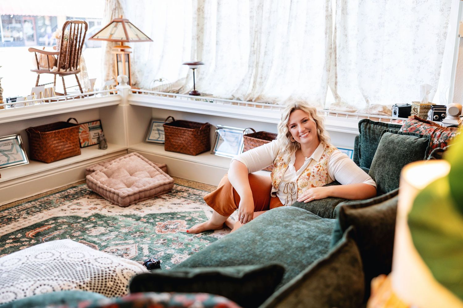 Smiling blonde woman relaxing on a green velvet sofa in a cozy, eclectic boutique shop with vintage decor.