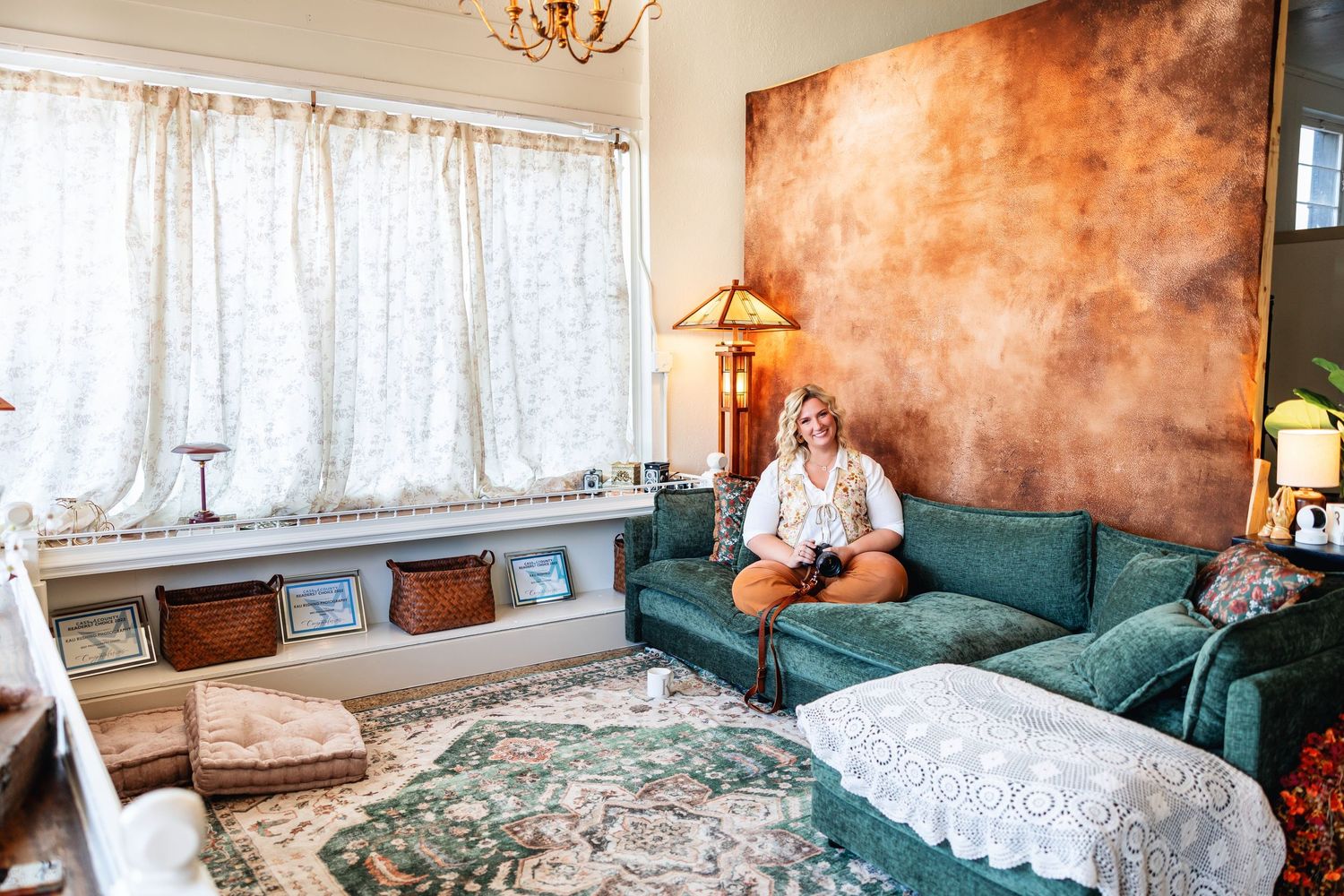 Blonde woman sitting on teal sofa in eclectic vintage studio with copper accent wall and lace curtains. Family photographer Texarkana TX