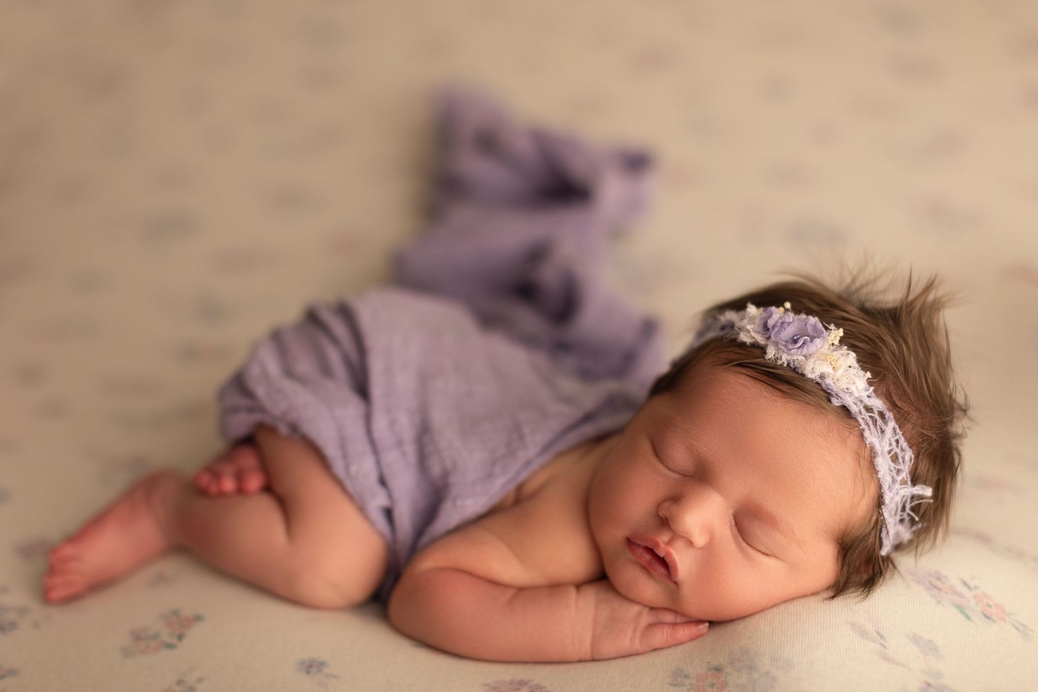Sleeping newborn baby girl wrapped in purple swaddle with floral headband on soft floral blanket.