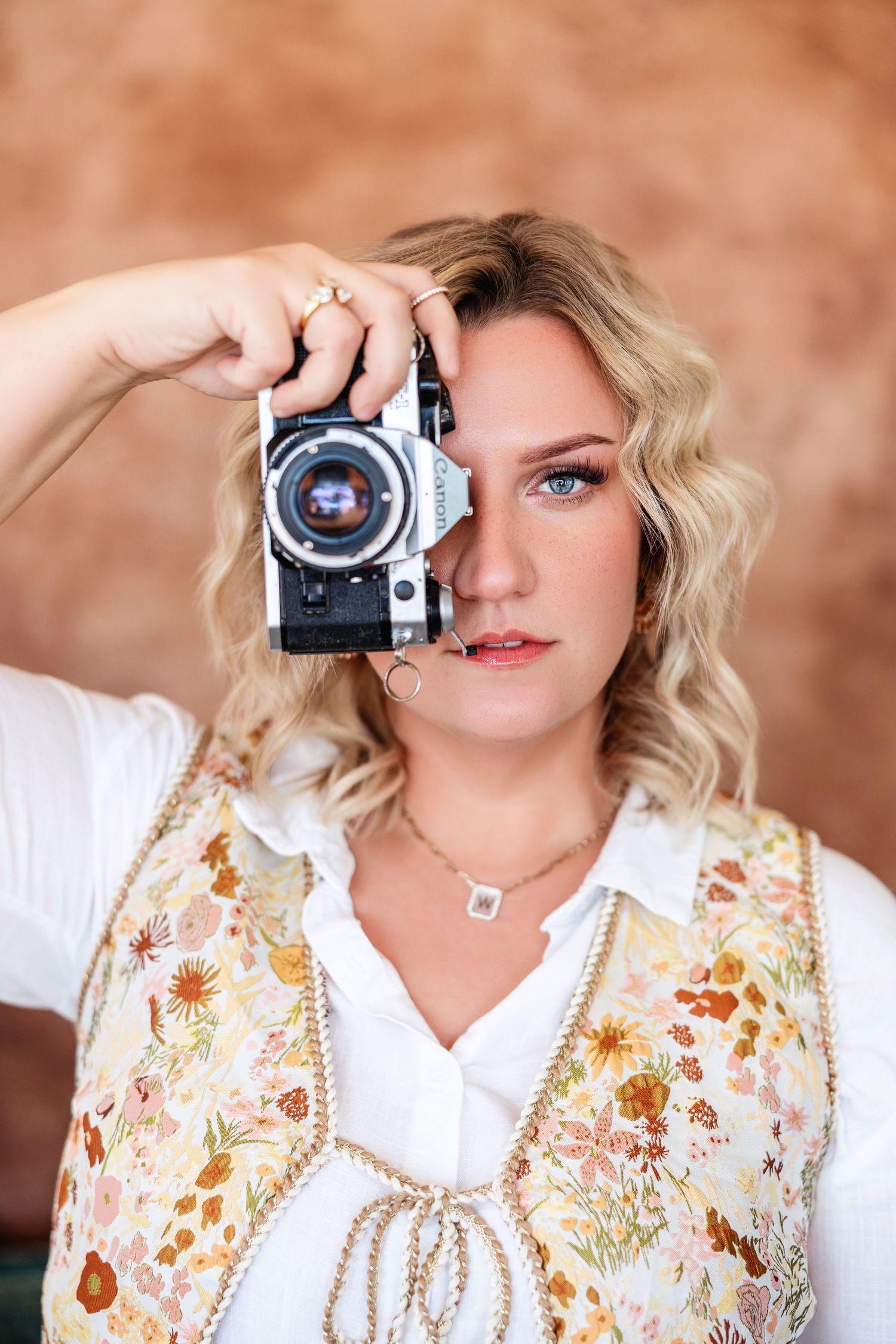 Blonde female photographer holding vintage film camera up to eye wearing floral vest in studio setting. Newborn photographer Texarkana TX