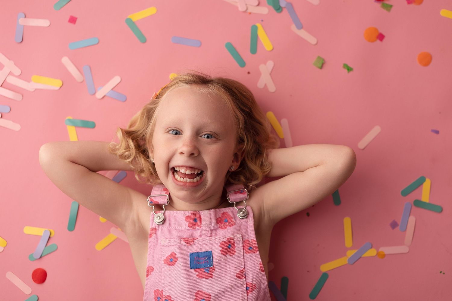 Joyful young girl with curly hair in pink overalls laughing against a pink background with colorful confetti.