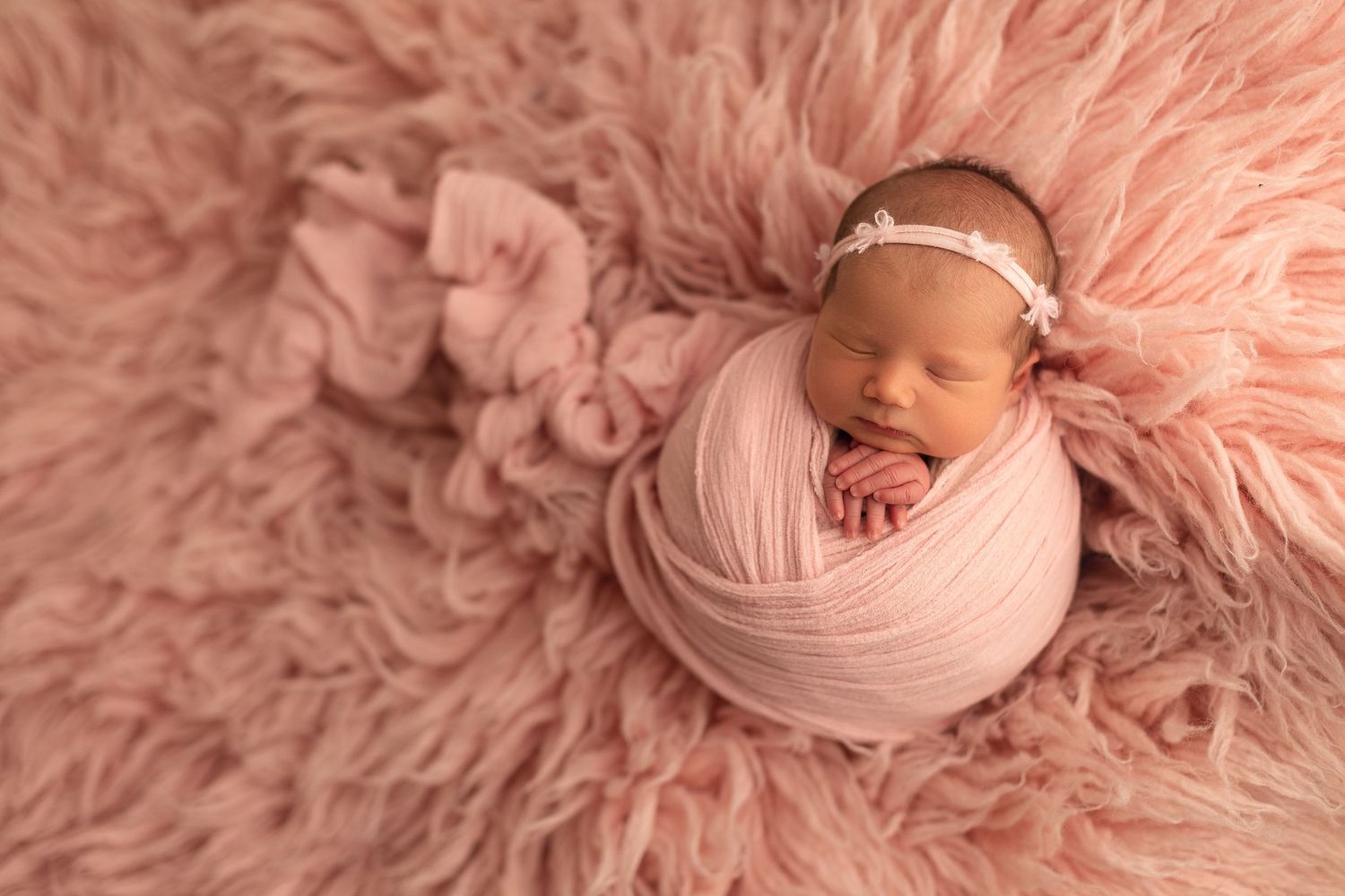 Newborn baby girl wrapped in pink swaddle on fluffy pink fur, wearing floral headband.